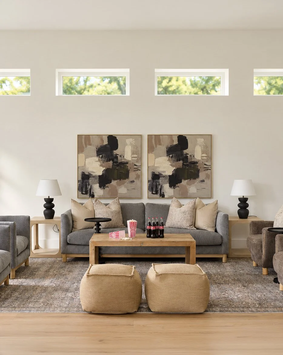 Living room with a gray sofa, abstract paintings, side tables with lamps, a wooden coffee table with soda bottles and popcorn, two beige poufs, and a patterned rug.