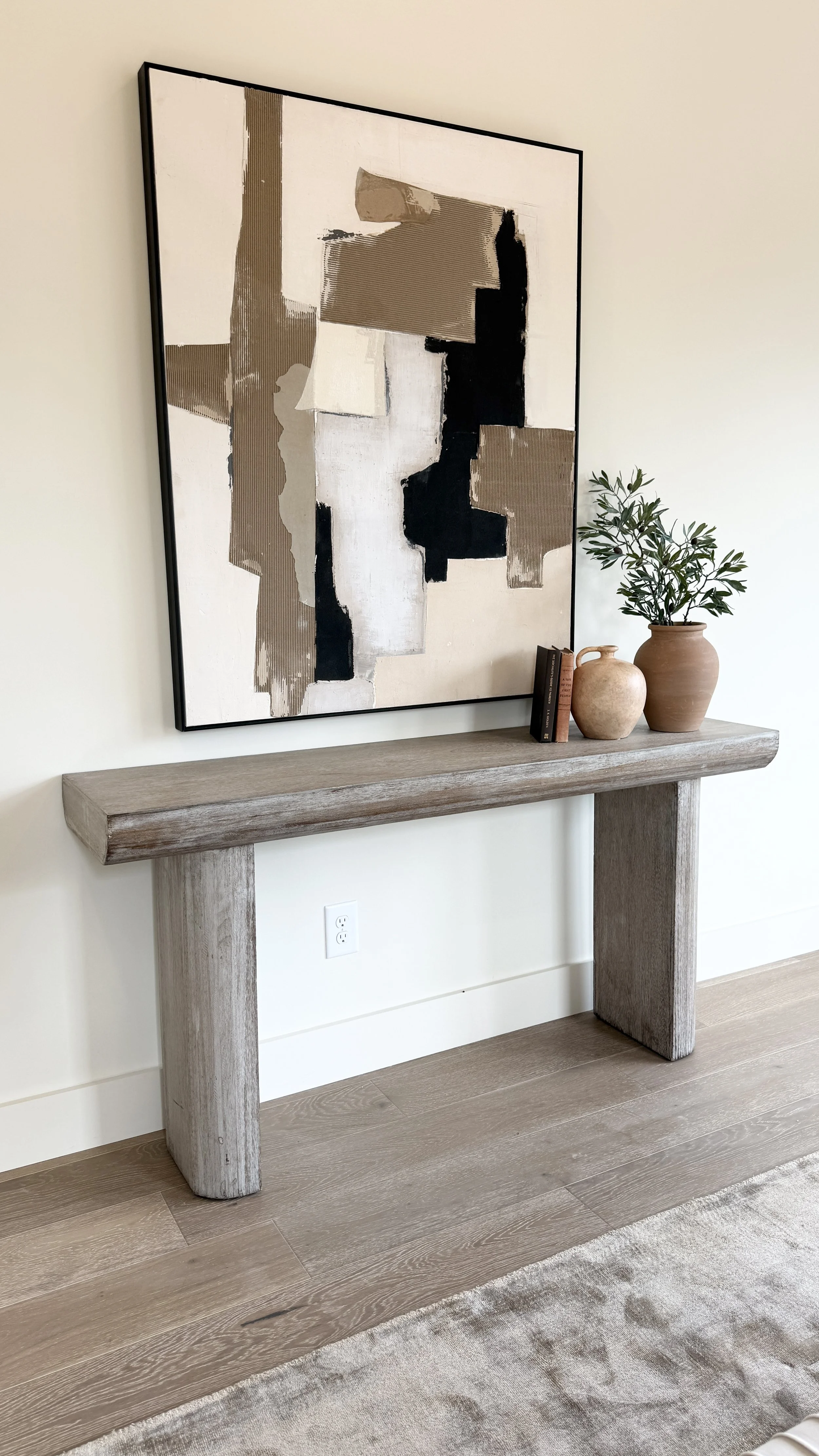 A framed abstract painting hangs above a rustic wooden console table decorated with potted plants and books, in a modern home interior.