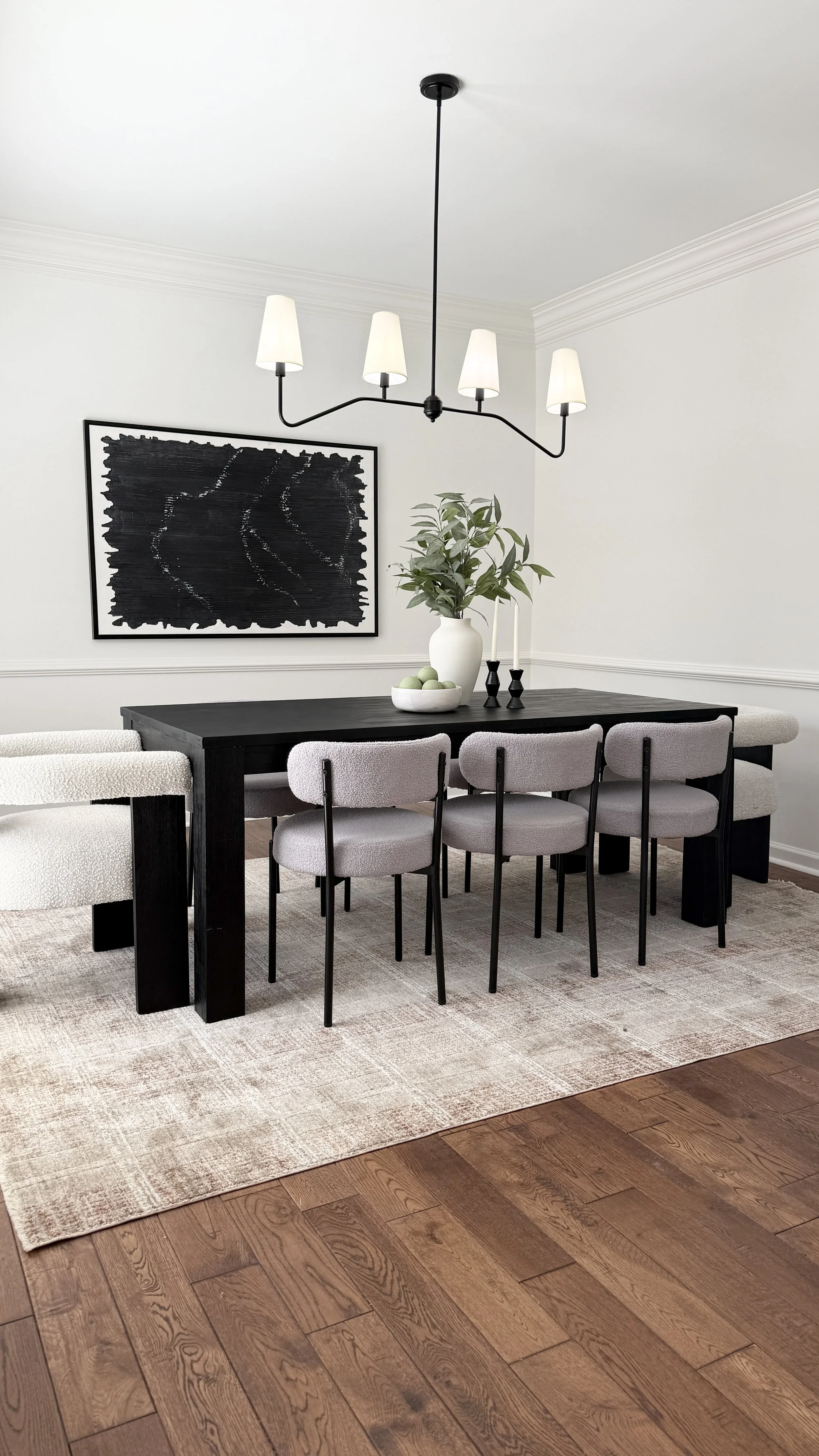 Interior dining room with a black table, white chairs, a beige rug, wall art, green plant, black candlesticks, and a black chandelier with white lampshades.