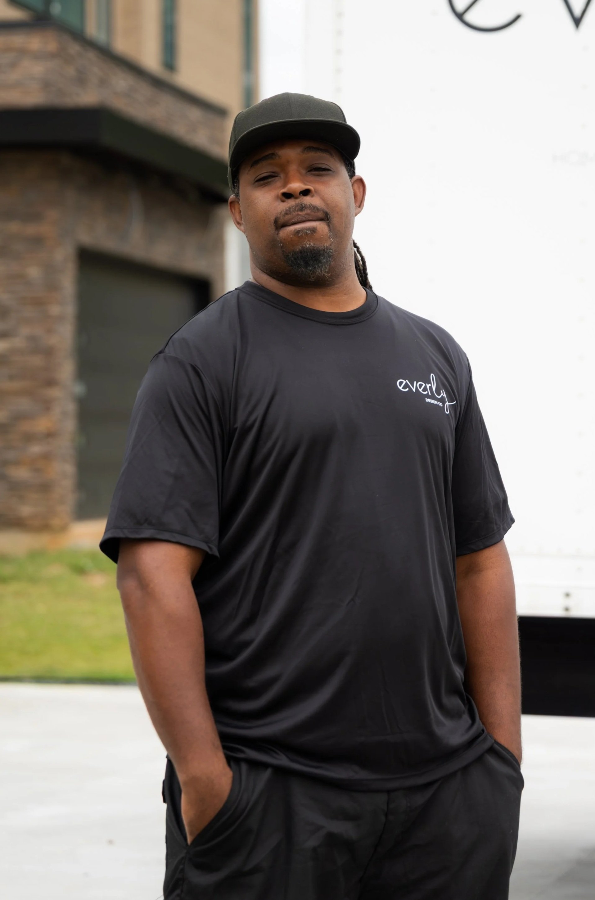 A man with dark hair and a beard standing outdoors, wearing a black t-shirt and a dark baseball cap, with a building and a white vehicle in the background.
