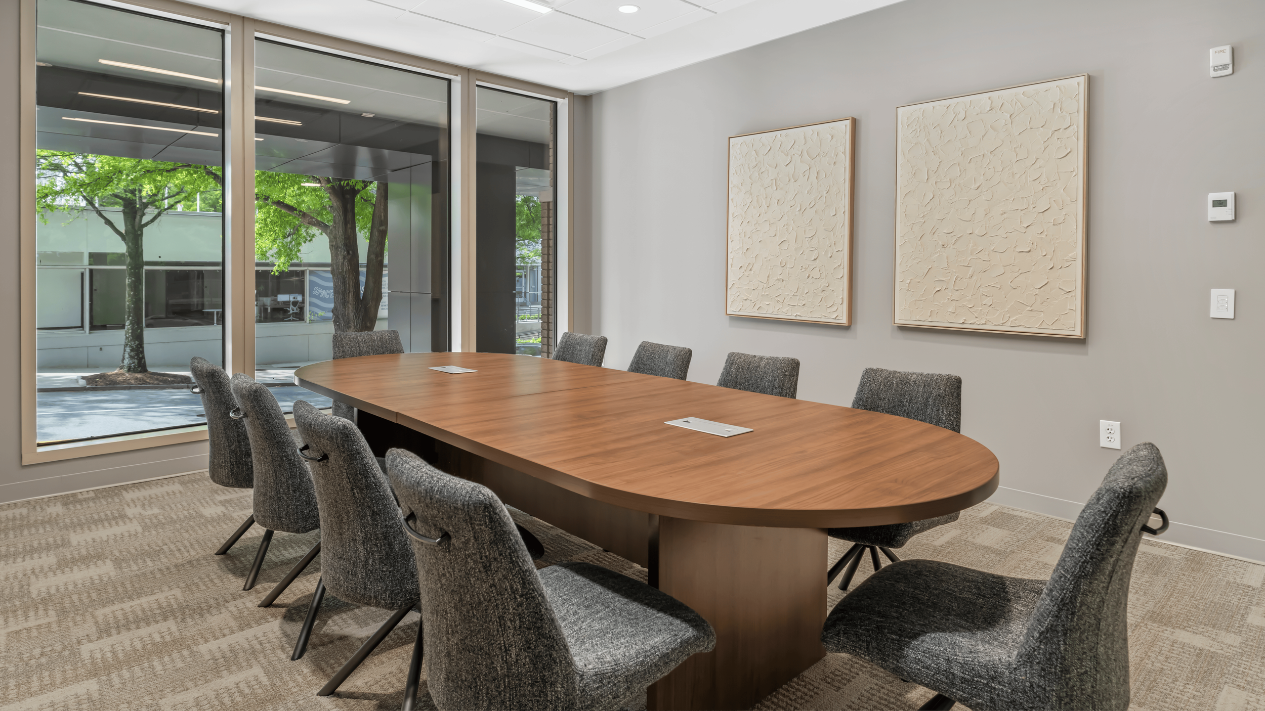 A modern conference room with a large oval wooden table surrounded by gray upholstered chairs. The room has large windows with views of trees outside and two abstract textured art pieces on the wall.