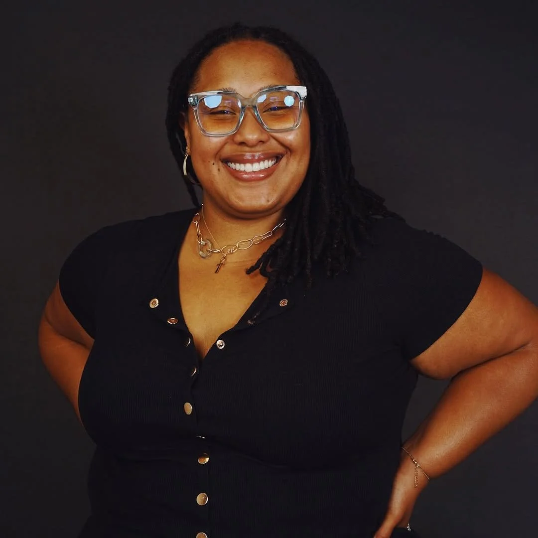 A smiling woman with dreadlocks, wearing glasses, a black top, and jewelry, posing with her hand on her hip against a dark background.