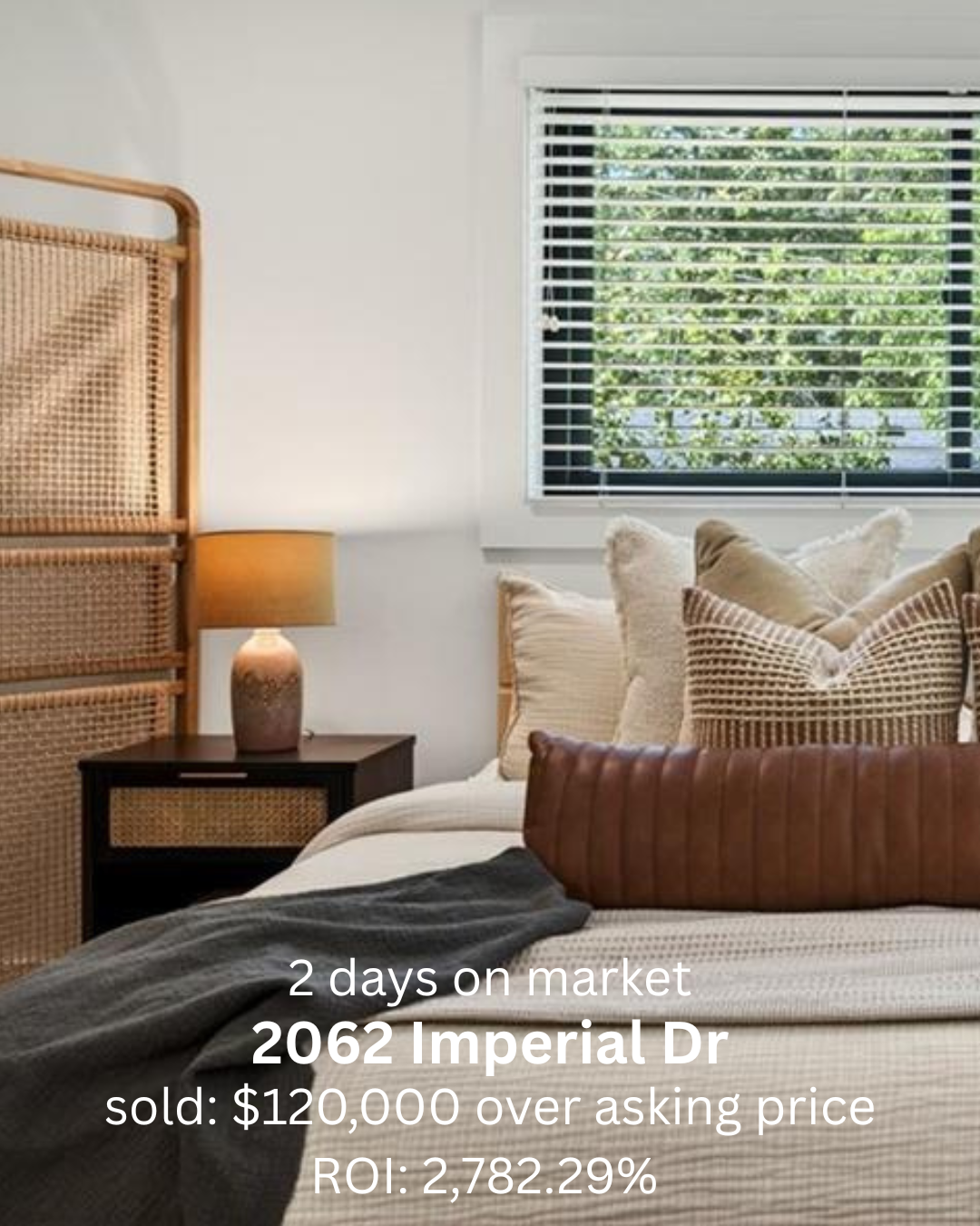 A bedroom with a bed, several pillows, a window with white blinds, a bedside table with a lamp, and a decorative rattan headboard.