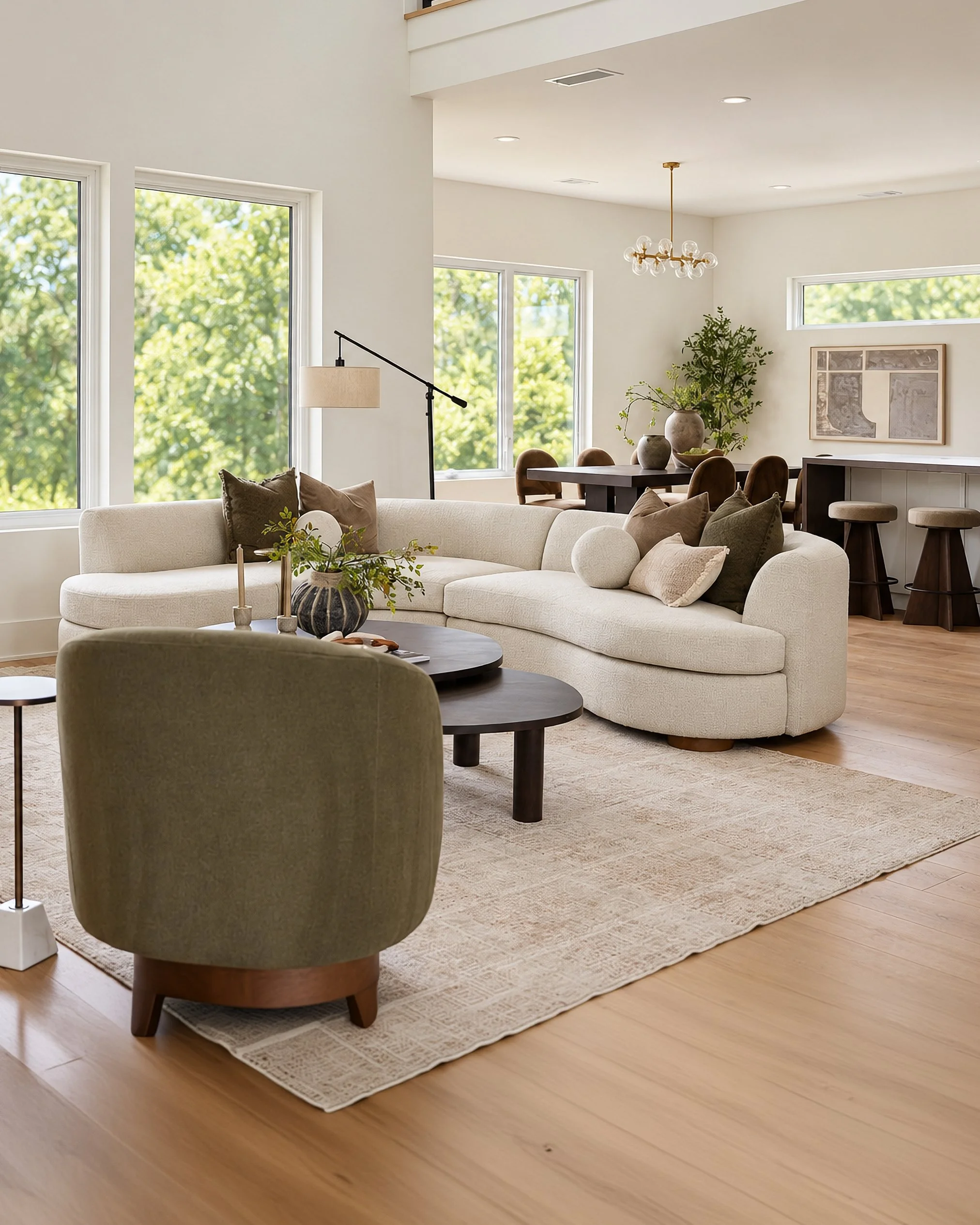 Sunlit living room with large windows, off-white sectional sofa with various pillows, a light-colored area rug, green armchair, round coffee table with decorative items, and modern decor including a floor lamp, artwork, and potted plants.