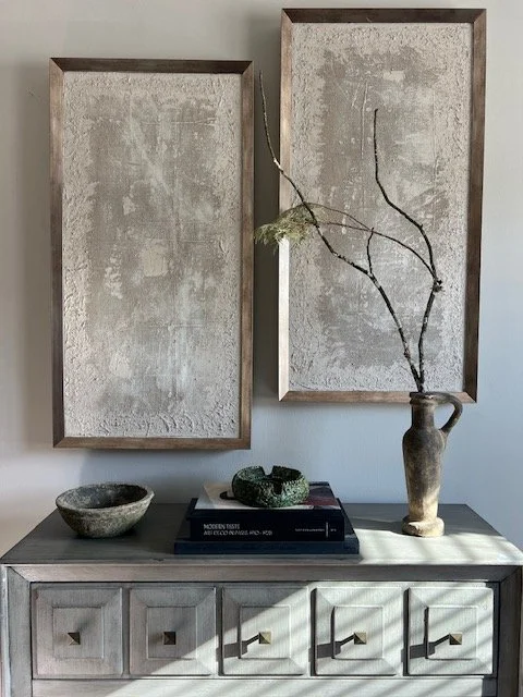 A wooden sideboard with five drawers and brass handles, topped with a large vase holding a branch with sparse leaves, a bowl, and stacked books. Two large, rectangular, textured art prints hang above the sideboard.