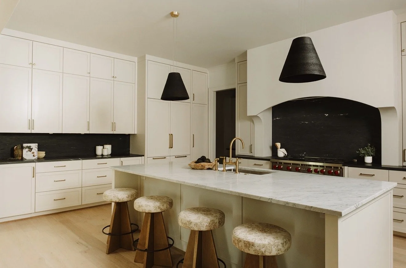 Modern kitchen with white cabinets, black countertops, a large white island with a marble top, three beige barstools, two black pendant lights, and a stove with a black backsplash.