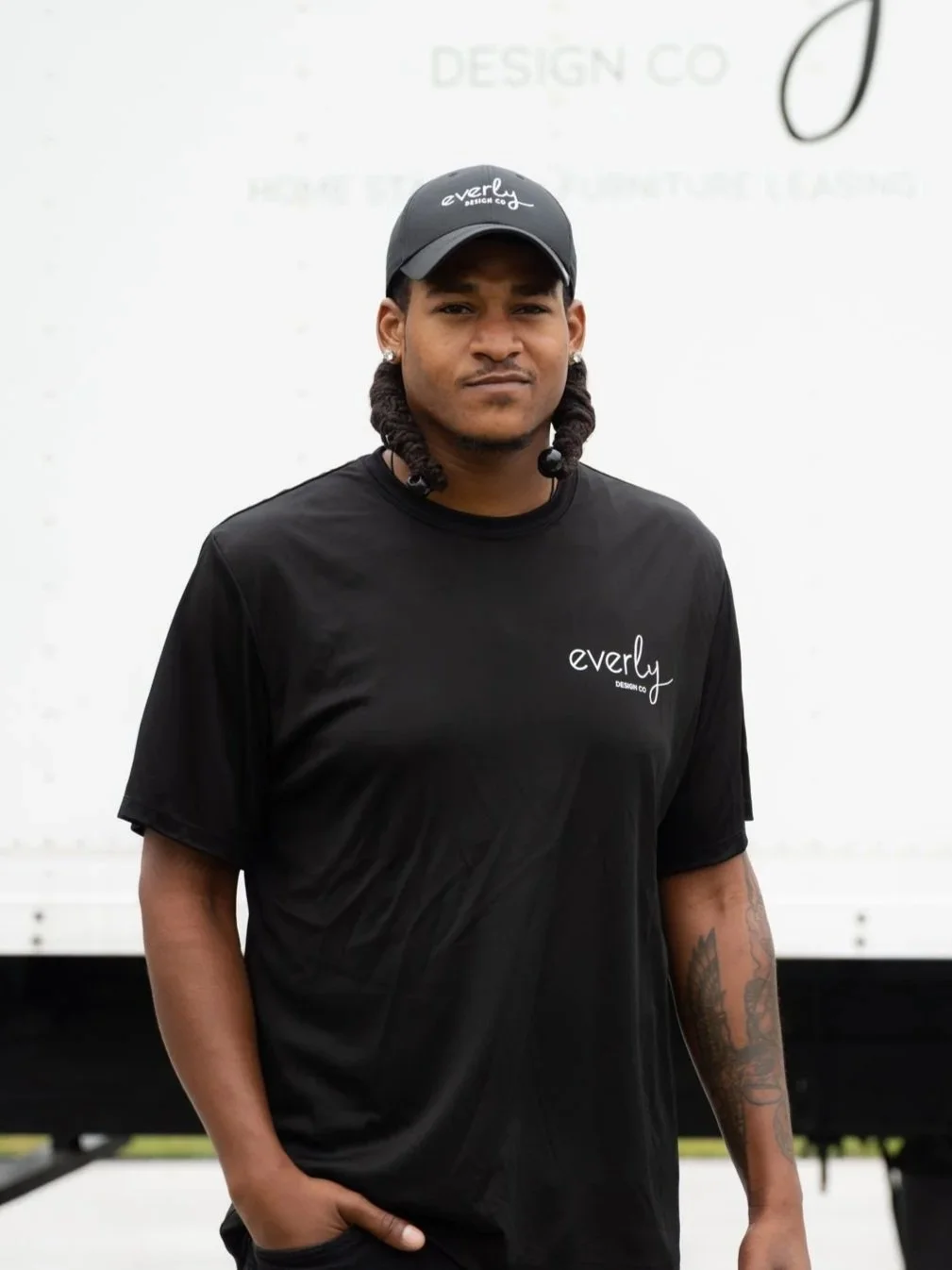 A man with medium skin tone and braided hair, wearing a black t-shirt and a black cap with the 'Everly Design Co' logo, stands outdoors in front of a white background with faint text.