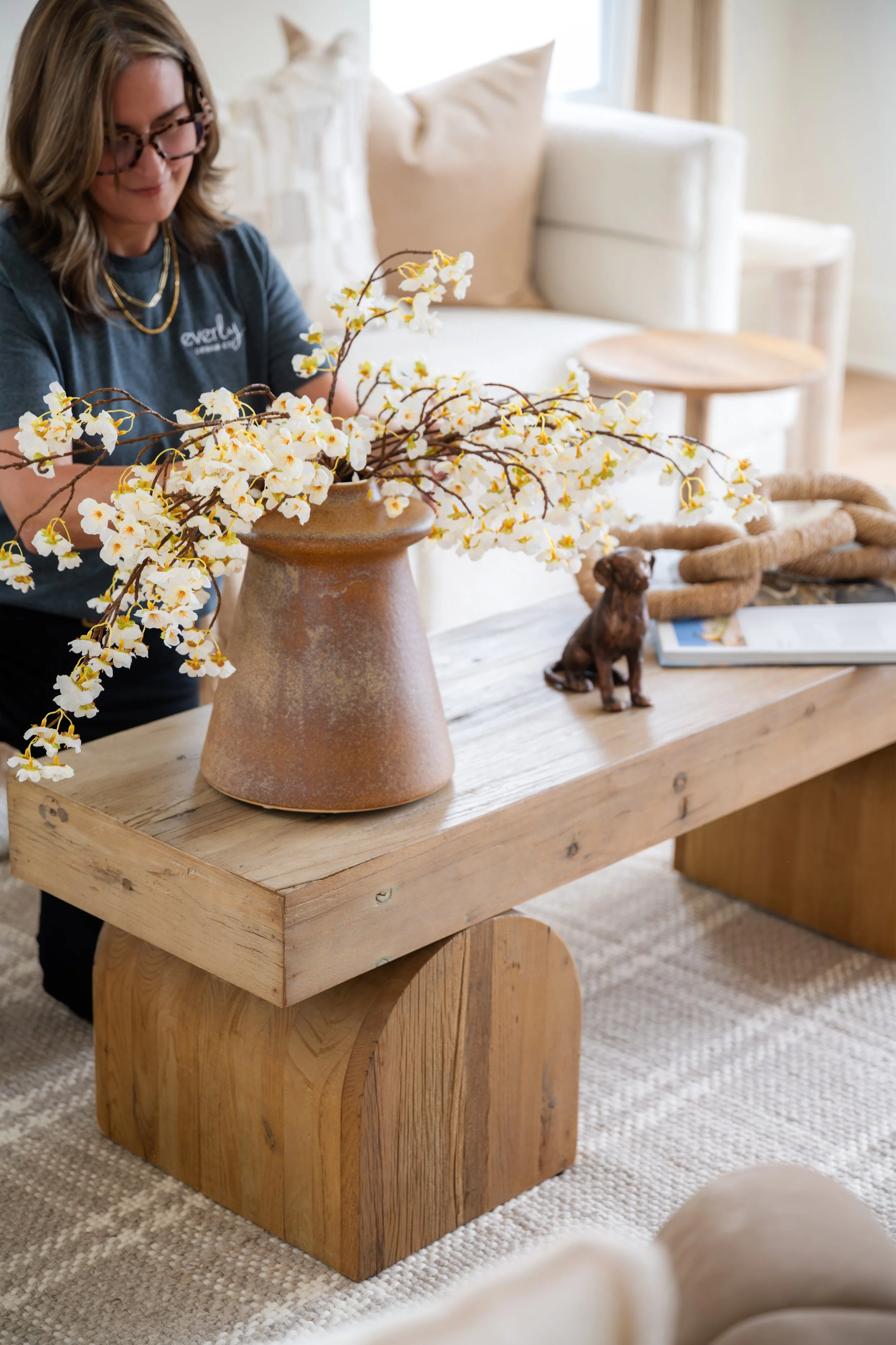 A woman with glasses and a dark t-shirt with the word 'evely' on it is decorating a large ceramic vase with white and yellow flowers on a wooden table. There is a small dog figurine, a rope, a magazine, and a picture frame on the table, in a well-lit room with a white couch and beige pillows.