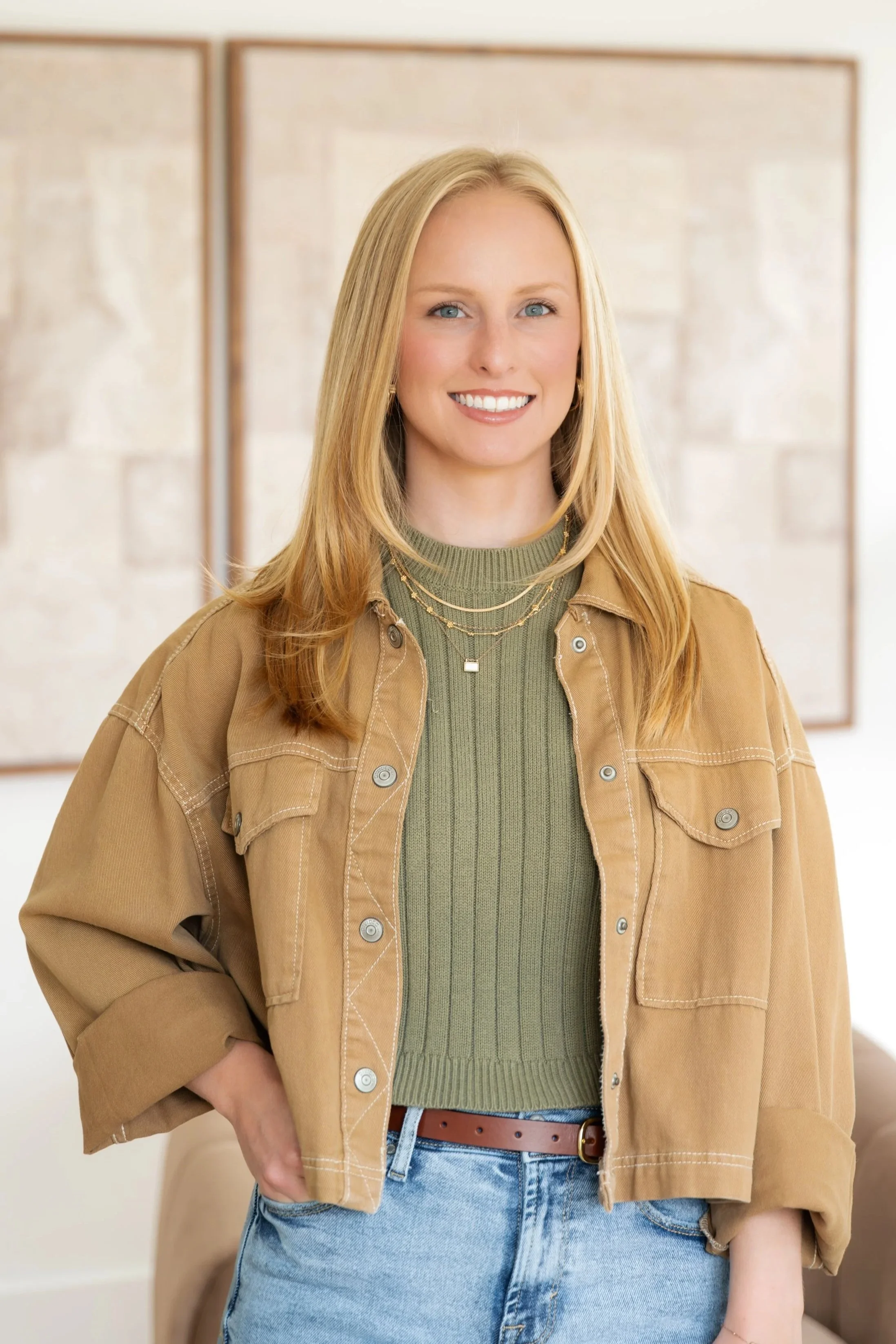 A woman with long blonde hair and blue eyes smiling, wearing a green sweater, tan jacket, and blue jeans, standing in front of a wall with framed artwork.