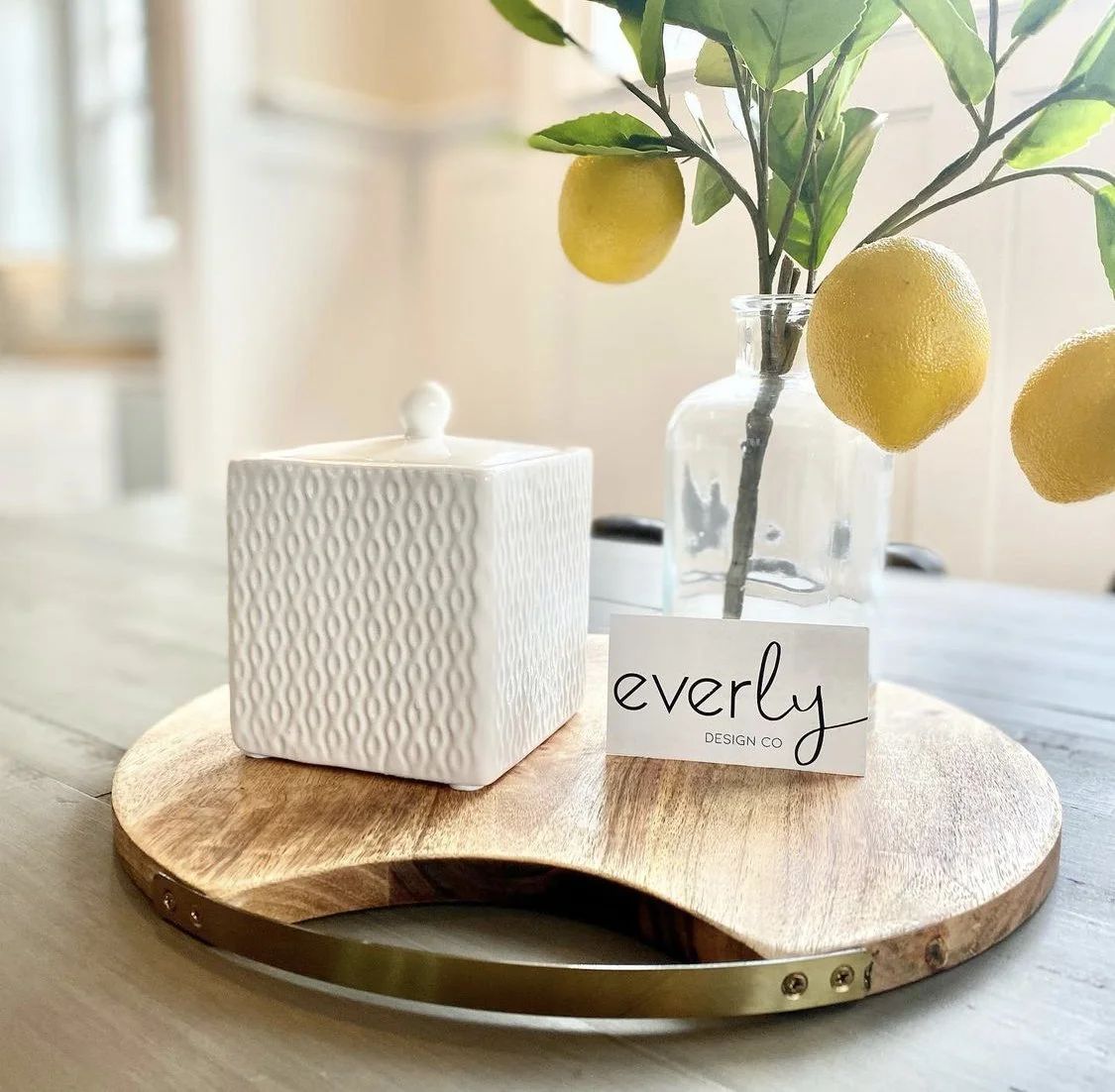 Decorative arrangement on a wooden tray with a white textured jar, a glass vase with lemon branches, and a business card reading 'everly DESIGN CO'.