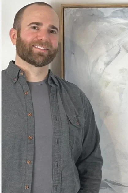 A man with a beard and short hair smiling, wearing a gray shirt over a darker gray t-shirt, standing indoors next to a framed abstract painting.