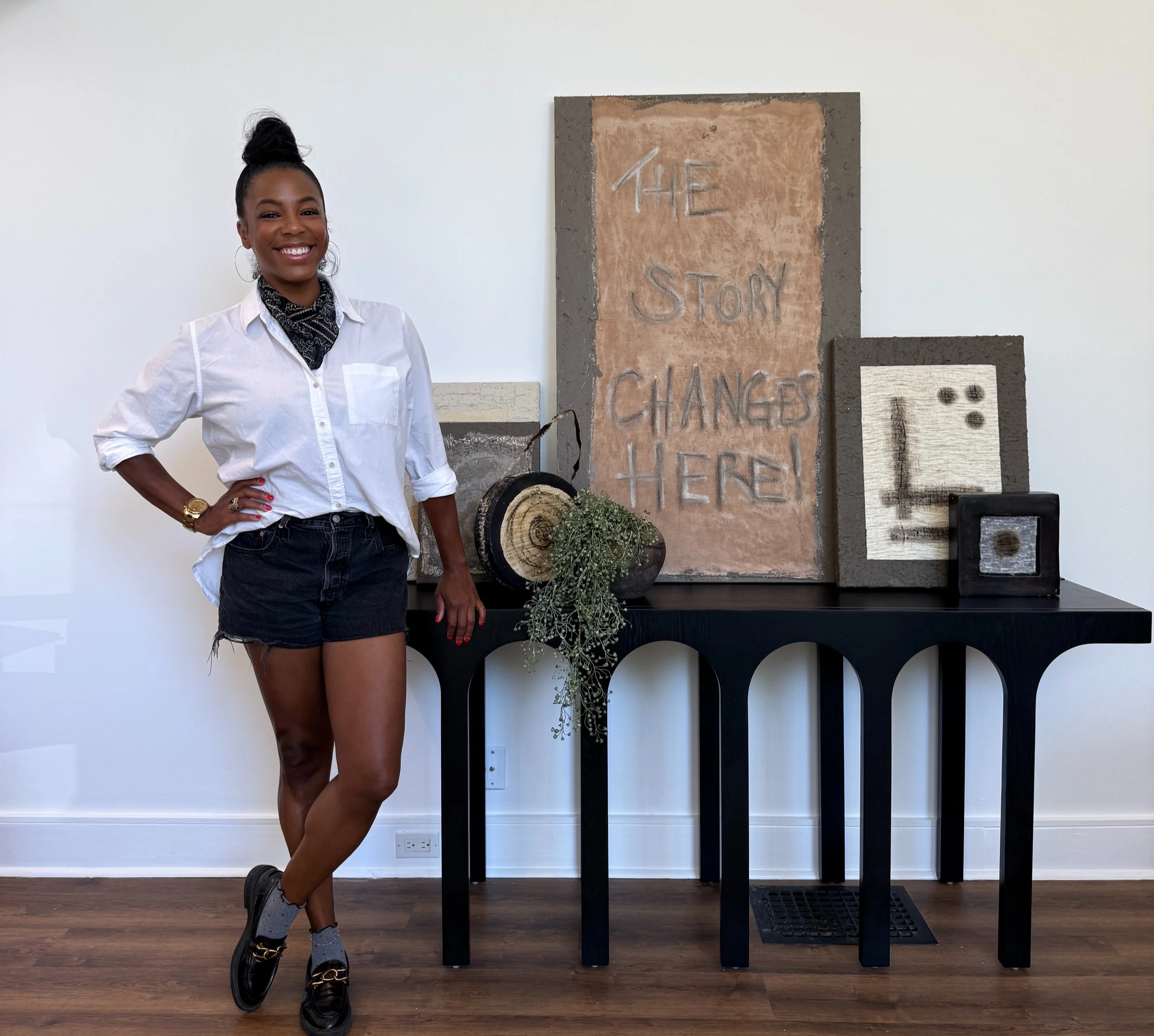 A woman with dark skin and hair in a high bun, wearing a white shirt, a black bandana around her neck, black shorts, and sneakers, stands in front of a display with a large, textured, rectangular artwork and smaller pieces, on a wooden floor with a plain white wall and electrical outlets in the background.
