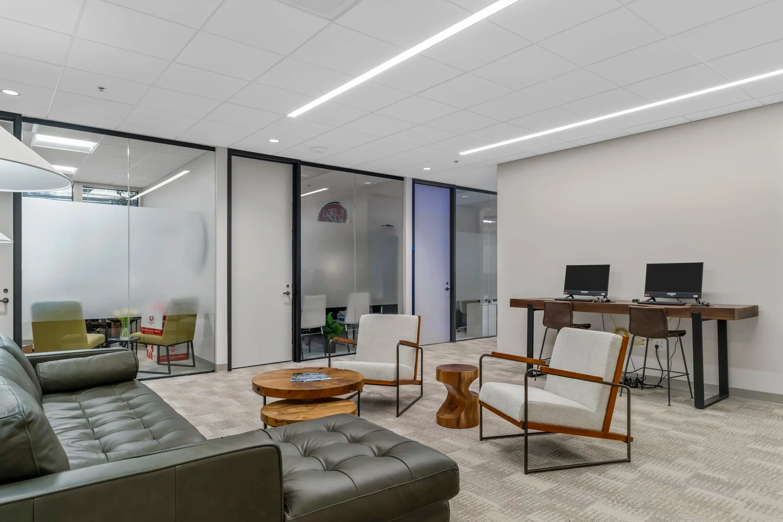 Modern office lounge with black leather sofa, white armchairs, wooden side tables, computer stations, glass partitions, and ceiling lights.