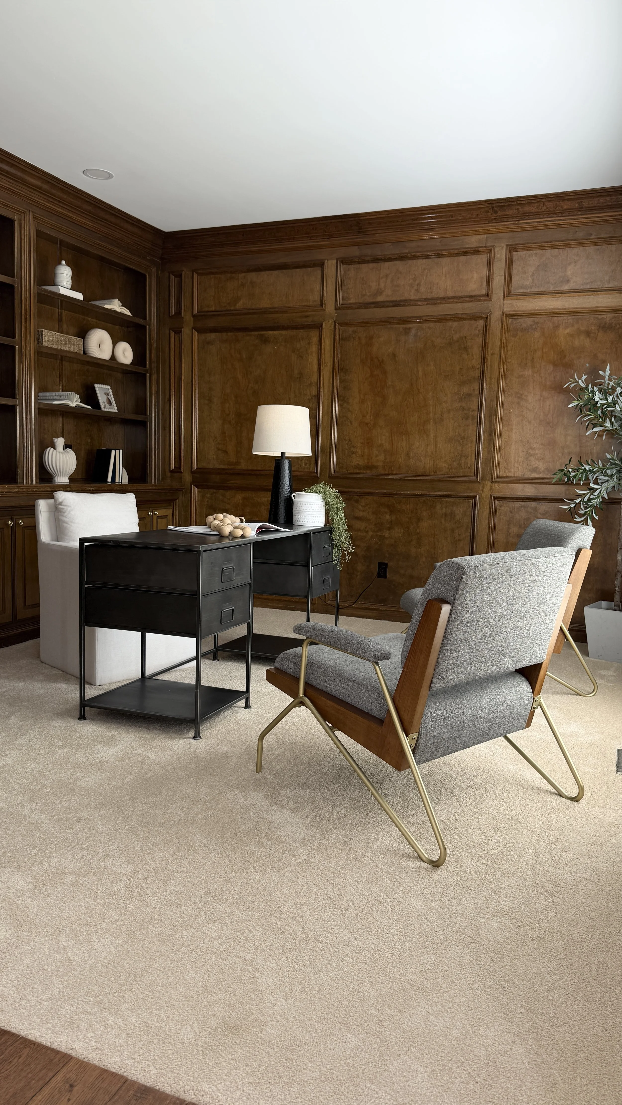 Living room with wood-paneled walls, a black side table, a white armchair behind it, a grey armchair, beige carpet, green potted plant, and decorative items on shelves.