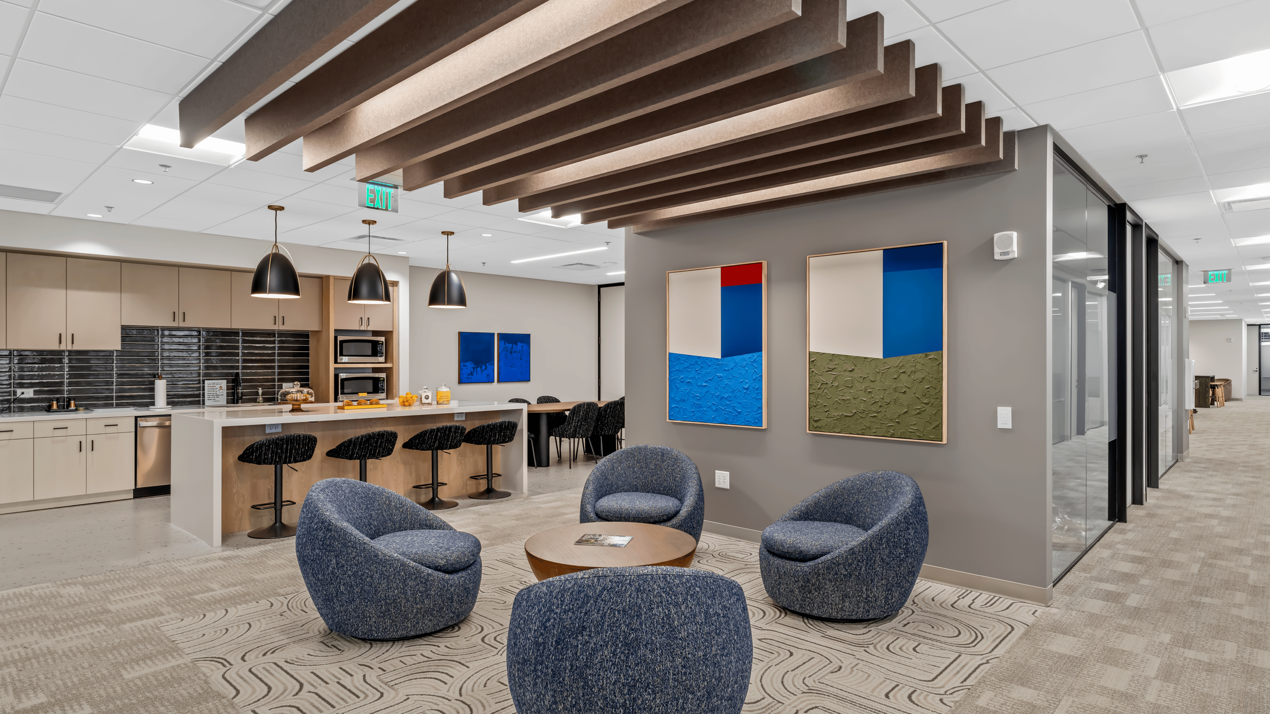 An office kitchen and lounge area with a breakfast bar, black barstools, blue armchairs around a small round table, and abstract art on the wall.