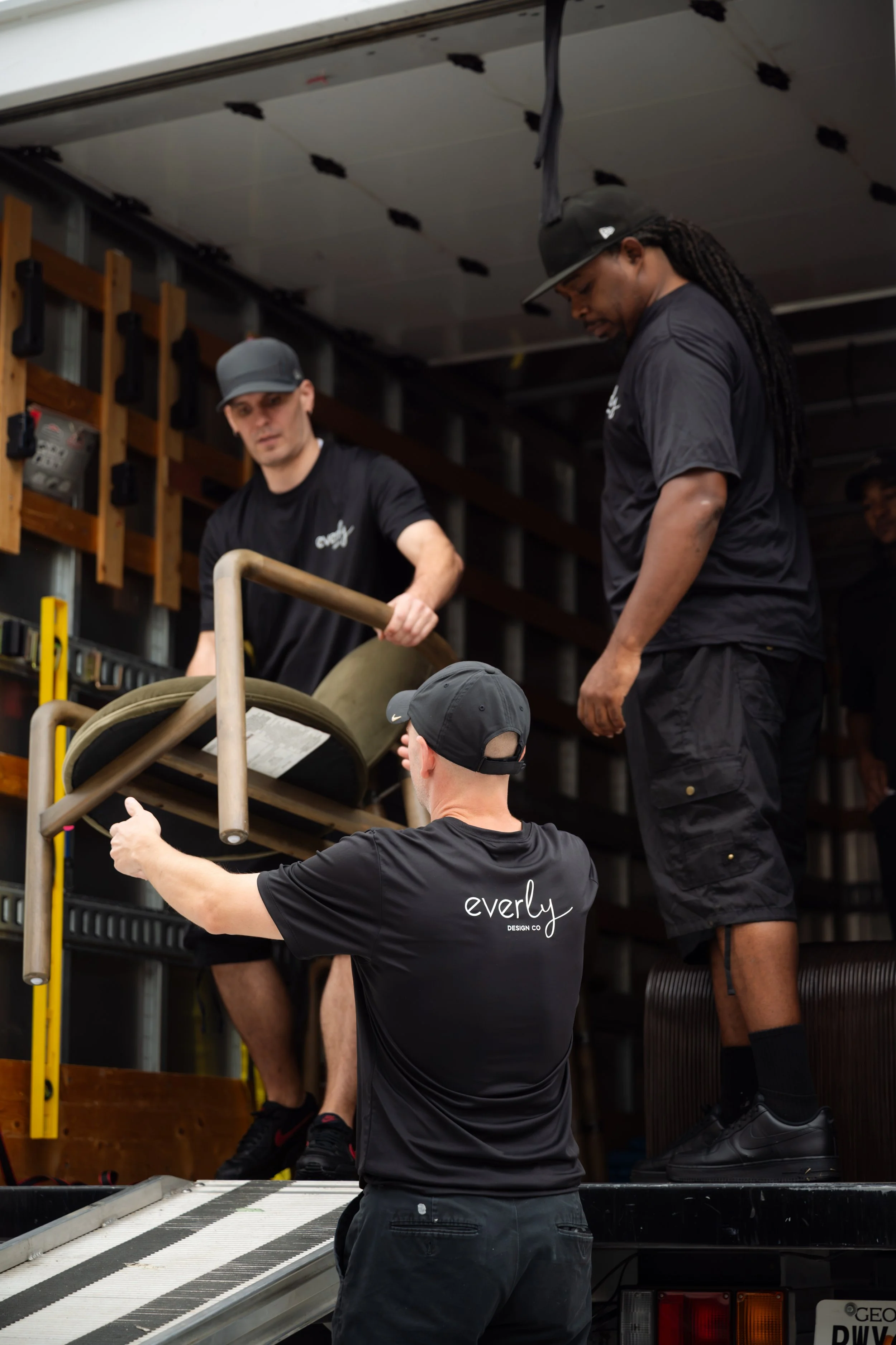 Three men wearing black shirts and hats working together around a large equipment or machinery in a workshop or garage with tools and equipment on the shelves behind them.