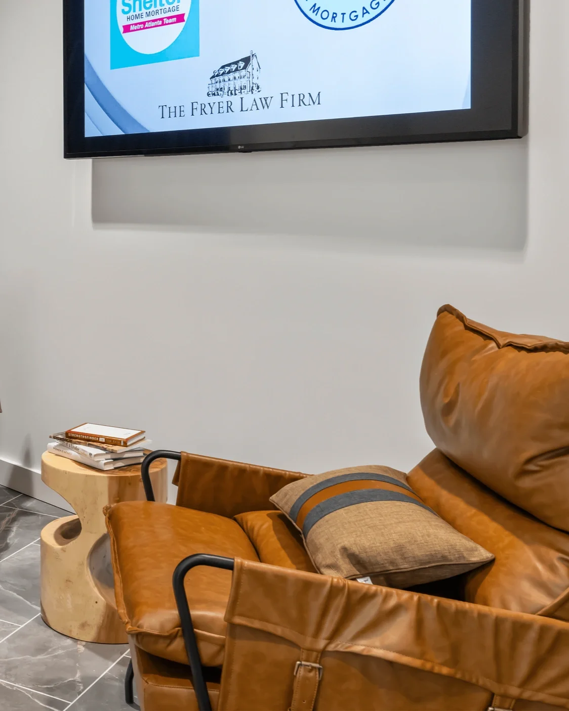 A leather sofa with a decorative pillow, a small wooden side table with books, and a wall-mounted television displaying various company logos in a modern office waiting area.