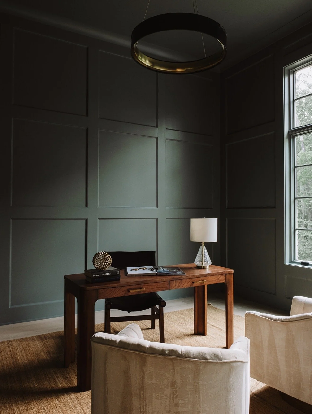 A home office with dark green paneled walls, a wooden desk, a white table lamp on a glass base, a black chair, a beige armchair, a window with natural light, a small decorative sphere, and a few books and documents on the desk.