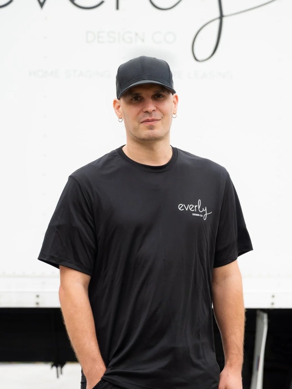 A man wearing a black cap and black T-shirt with the word "everly" on it, standing against a white background with partial visible text.