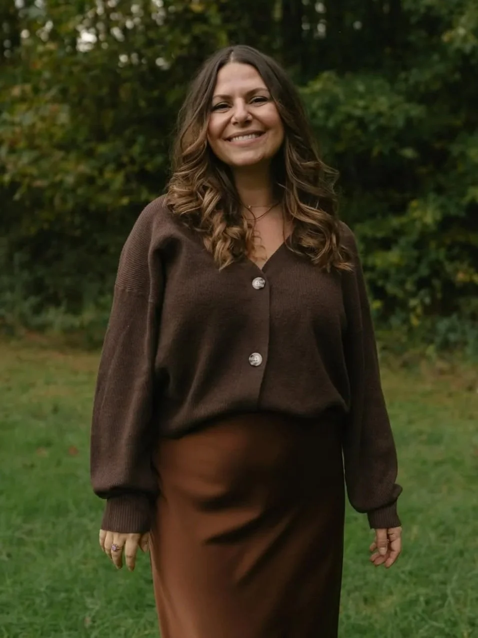 A woman with long wavy brown hair, smiling, wearing a brown cardigan and matching skirt, standing outdoors in a park with green trees in the background.