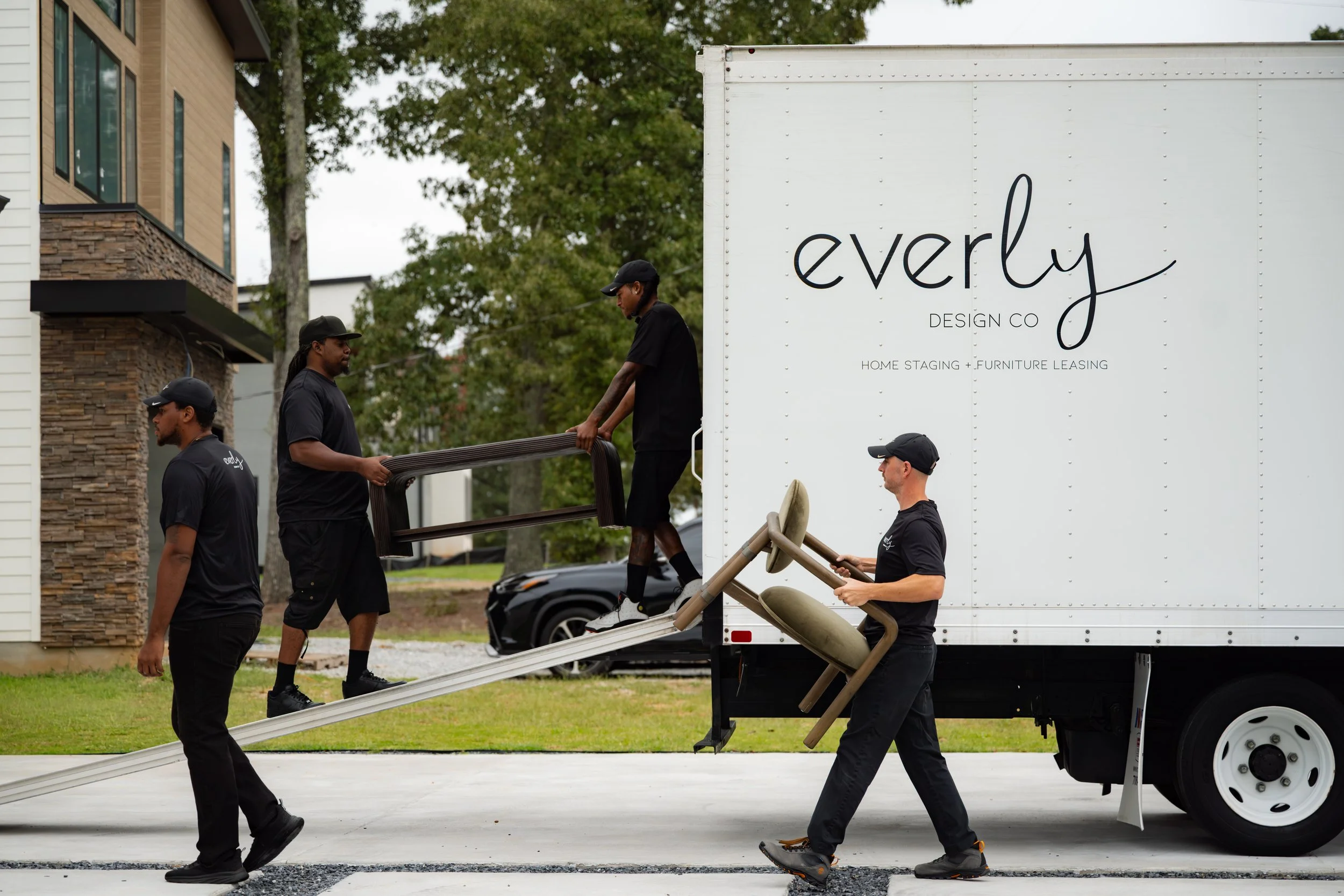 A group of five workers unloading furniture from a moving truck in a residential area during daytime. They are carrying a table and a window frame.