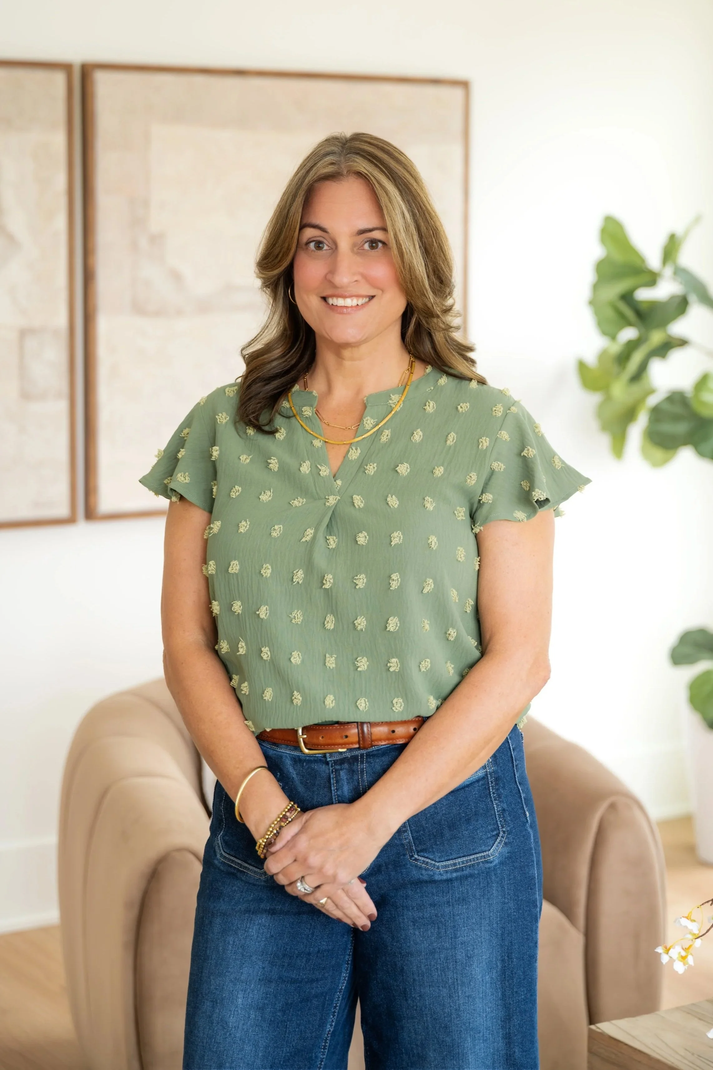 A woman with shoulder-length brown hair, wearing a green blouse with small yellow flowers, jeans, and jewelry, standing in a room with light-colored walls, a beige armchair, and a large green plant.