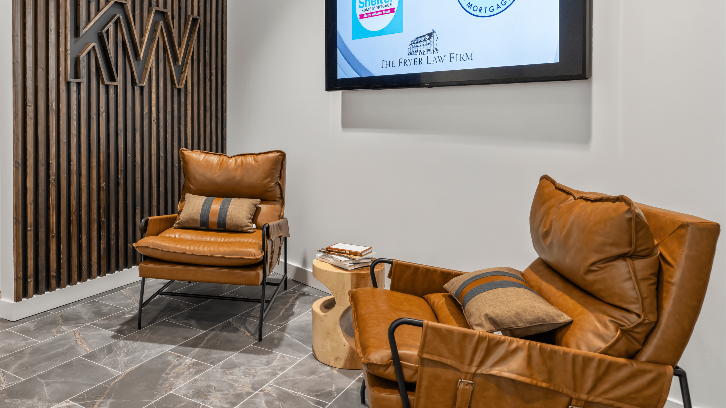 A waiting area with two brown leather chairs featuring black metal frames and matching cushions, a small wooden side table with books on it, a decorative wooden wall, and a mounted TV displaying logos of local businesses.