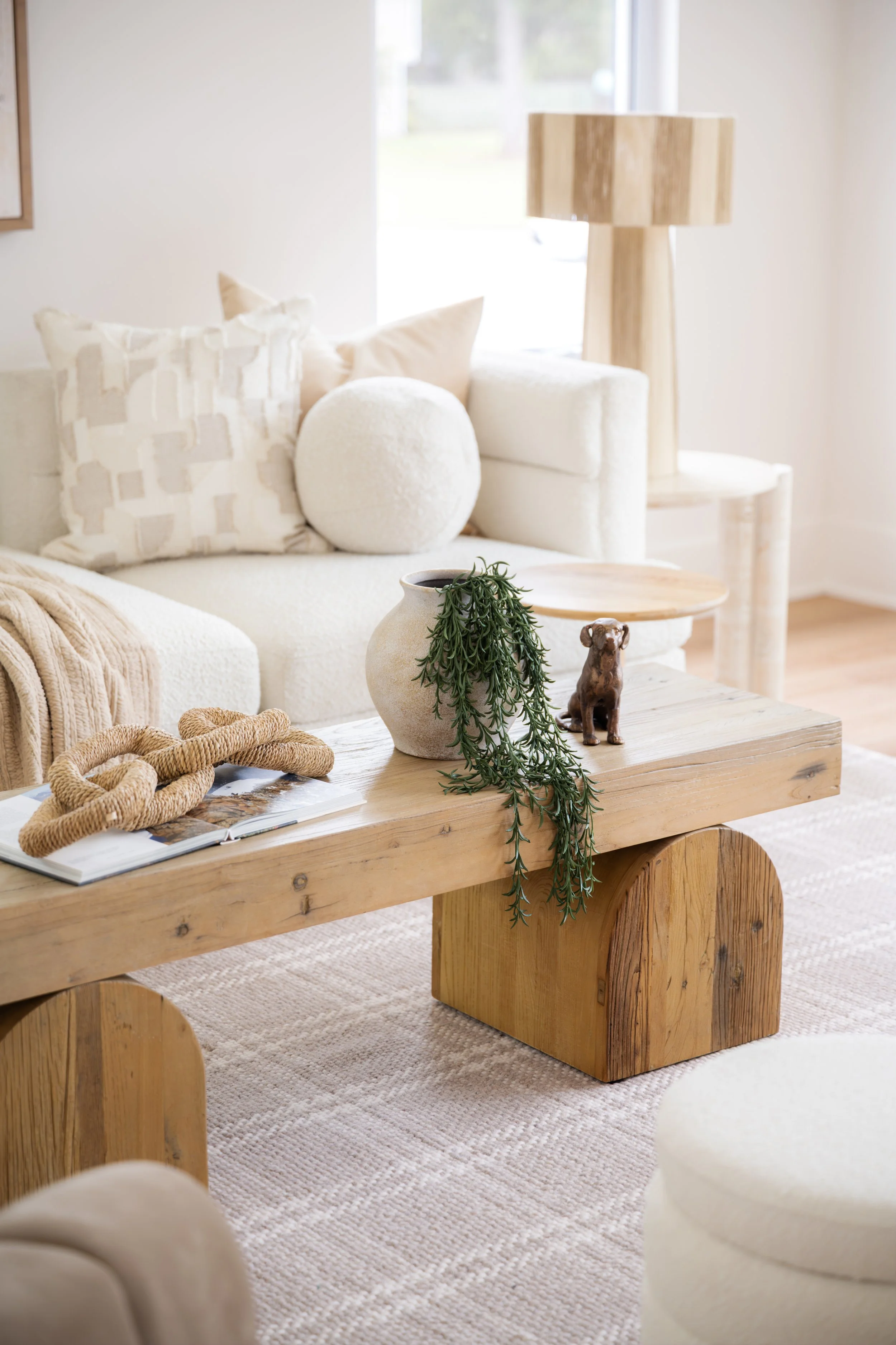 A living room with a neutral-colored sofa, decorative pillows, a wooden coffee table with a plant and a small dog figurine, and a wooden lamp, with natural light coming through a window.