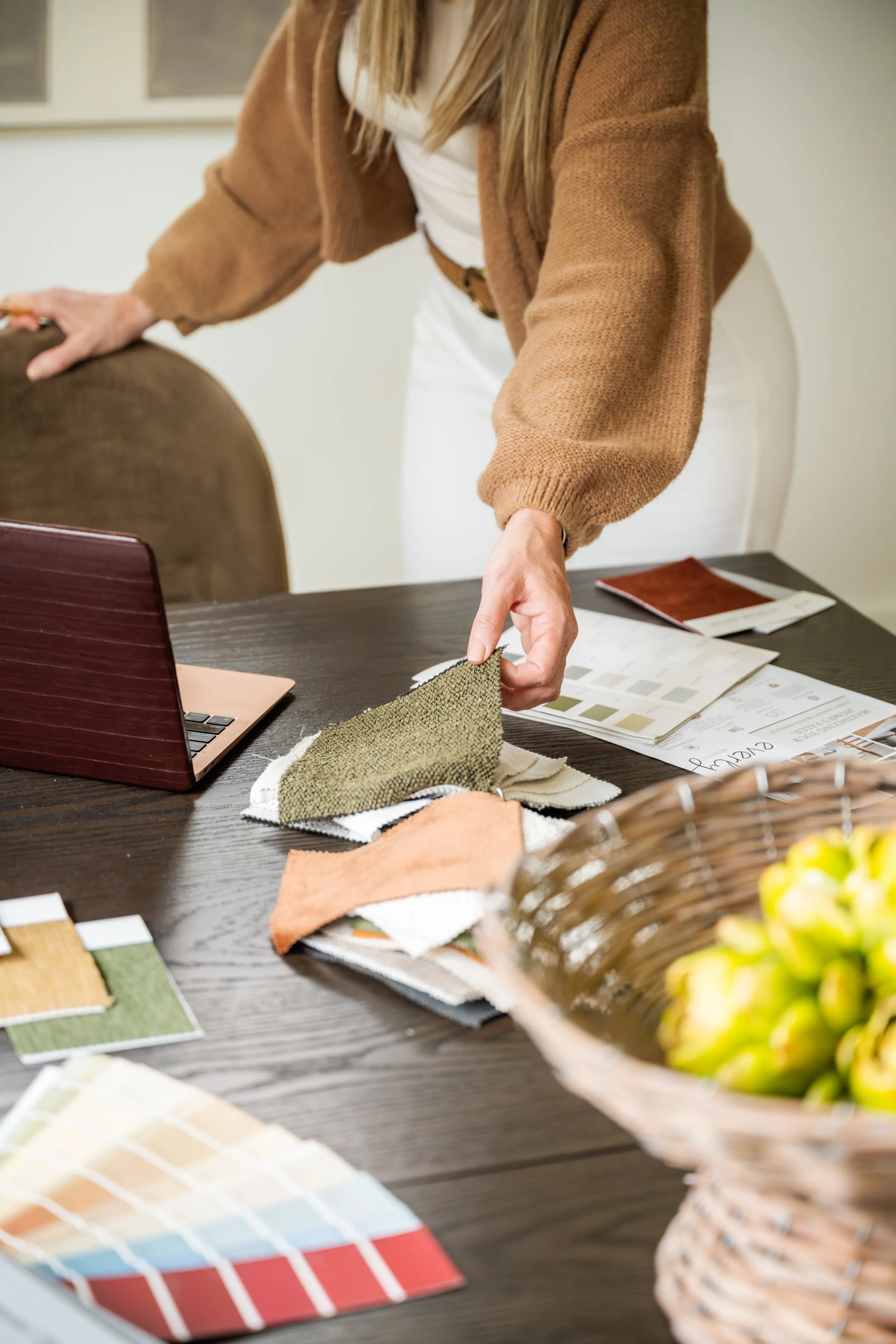 A woman is examining fabric swatches and color samples on a table, with design books, color palettes, and textiles spread out. There is a laptop and a basket of yellow flowers on the table.