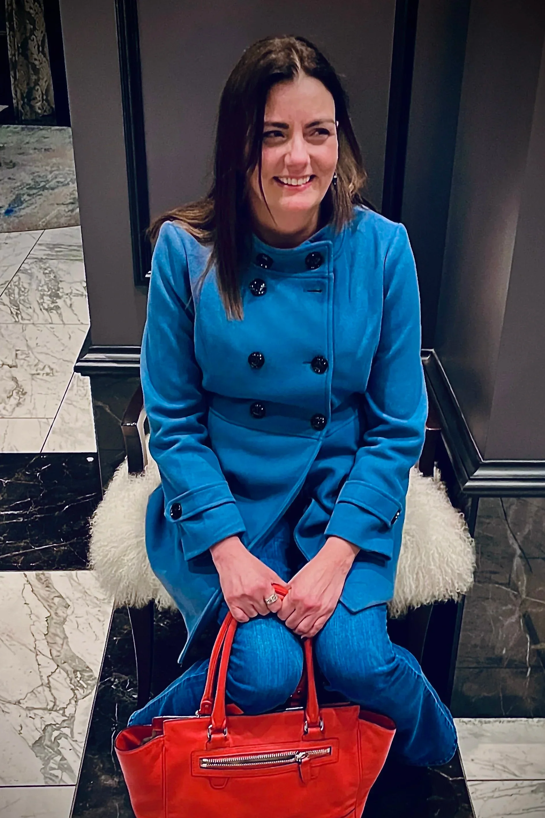 A woman with dark brown hair, wearing a blue jacket with black buttons, sitting on a chair with a white furry cushion, holding an orange handbag, smiling in an indoor setting with marble floors and dark walls.