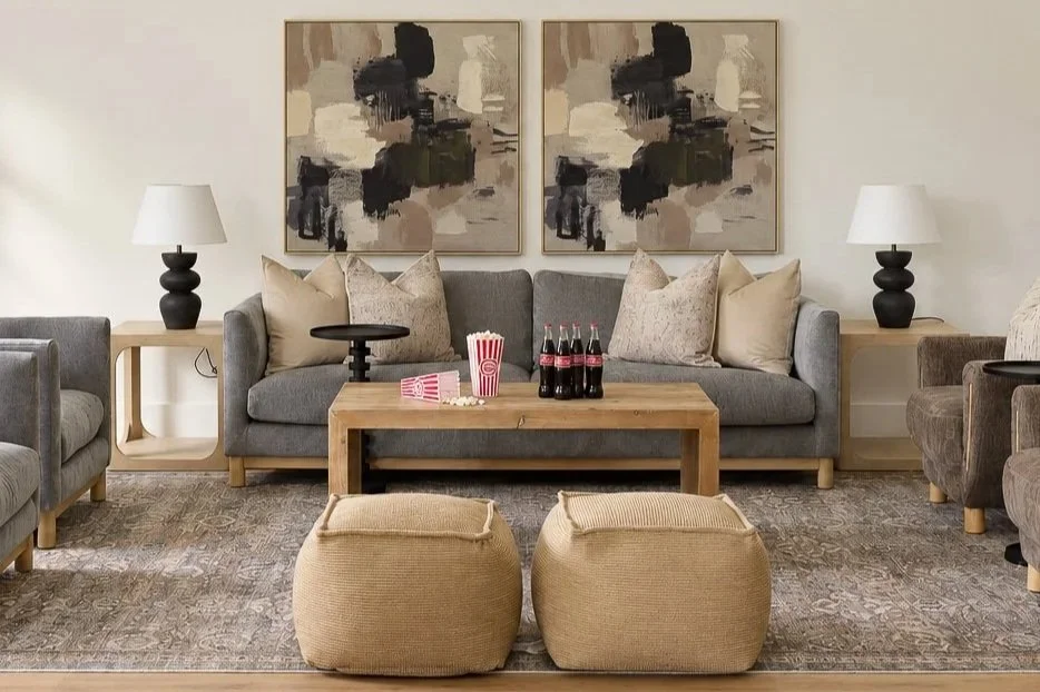 Living room with gray sofa, abstract black and beige wall art, two beige ottomans, side tables with lamps, and a wooden coffee table with soda bottles and popcorn containers.