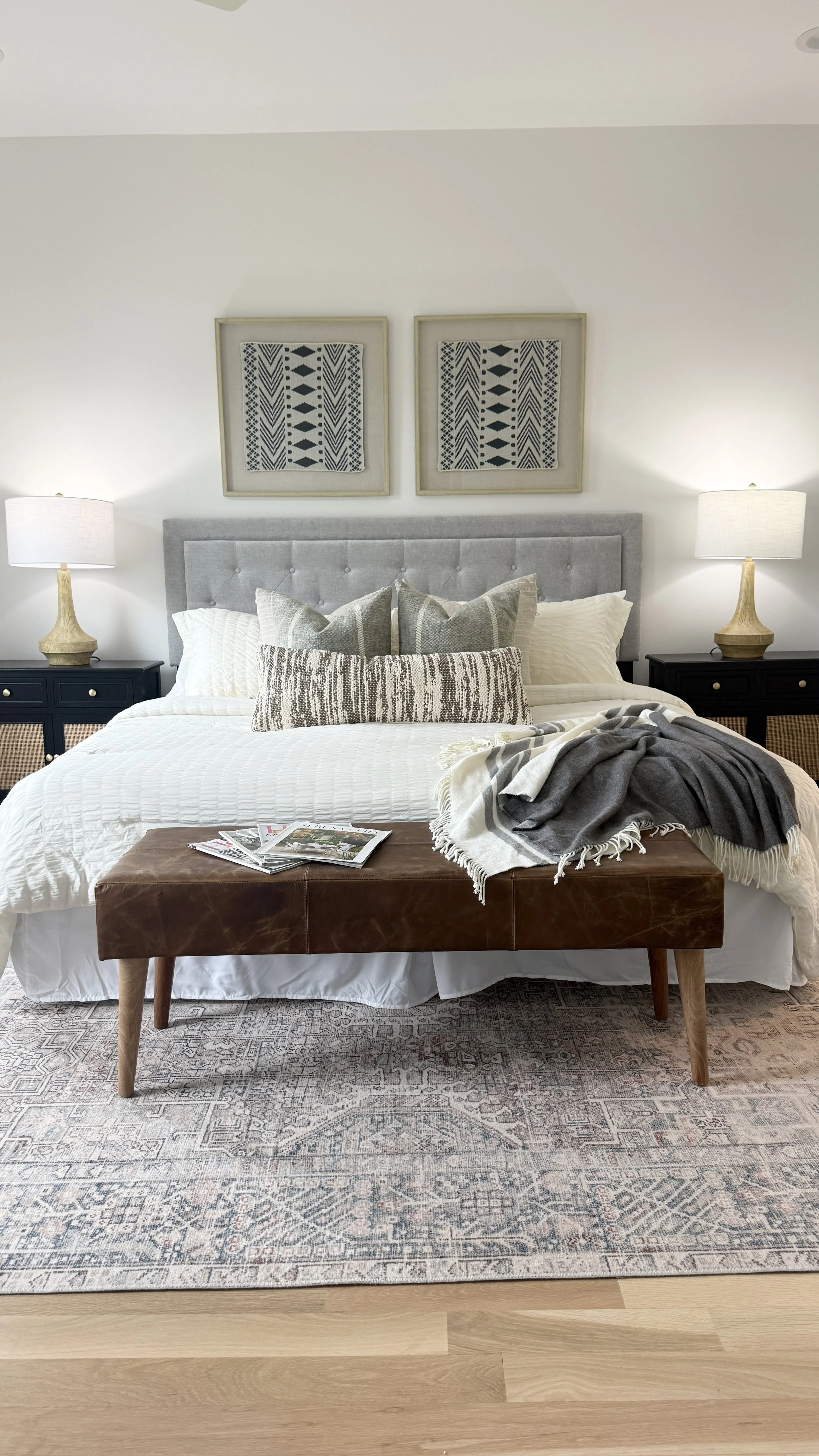 A cozy bedroom with a neatly made bed, gray upholstered headboard, and a mix of white, gray, and patterned pillows. Two black nightstands with beige lamps flank the bed. Above, two framed black and white geometric artworks hang on the white wall. A leather ottoman at the foot of the bed has magazines and a gray and white throw blanket draped over it. The room features a patterned rug on light hardwood flooring.