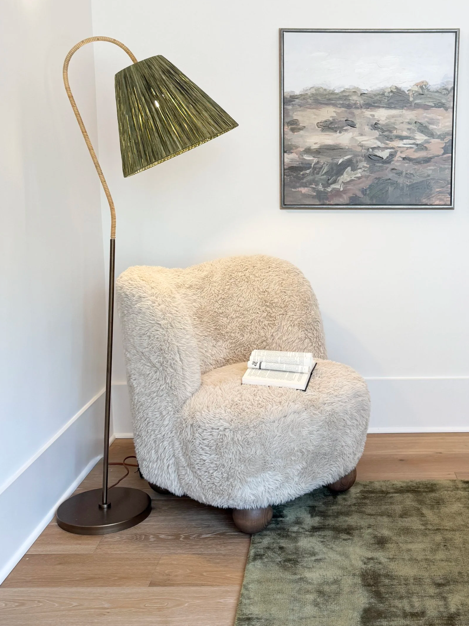 A cozy reading nook with a fluffy beige armchair, an open book on it, a tall floor lamp with a brass finish and a green lampshade, a light-colored wall with an abstract landscape painting, light wood flooring, and a muted green rug.
