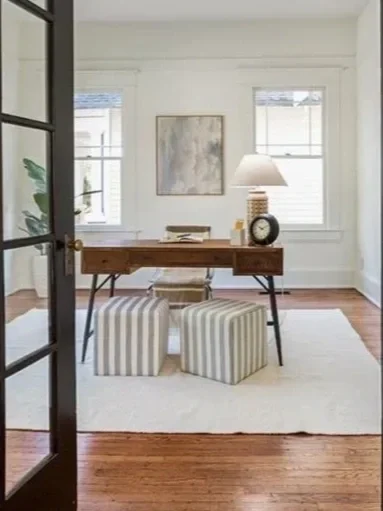 A well-lit home office with a wooden desk, two striped stools, a table lamp, a clock, and artwork on the wall.