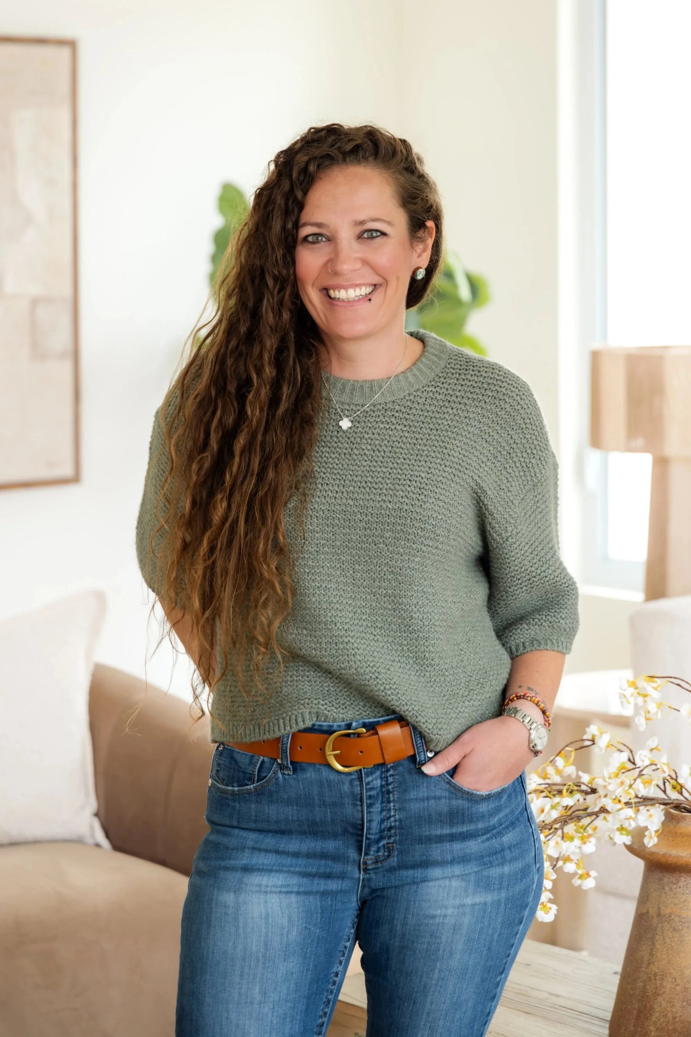 A woman with long, curly brown hair and blue eyes smiling, wearing a green knitted sweater, blue jeans, a brown belt, and jewelry, standing in a cozy living room with a beige sofa, white flowers, and a large window in the background.