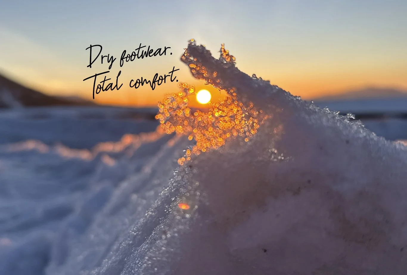 Close-up of a wave breaking on the beach with the sun setting in the background, and the text 'Dry footwear. Total comfort.' on the image.