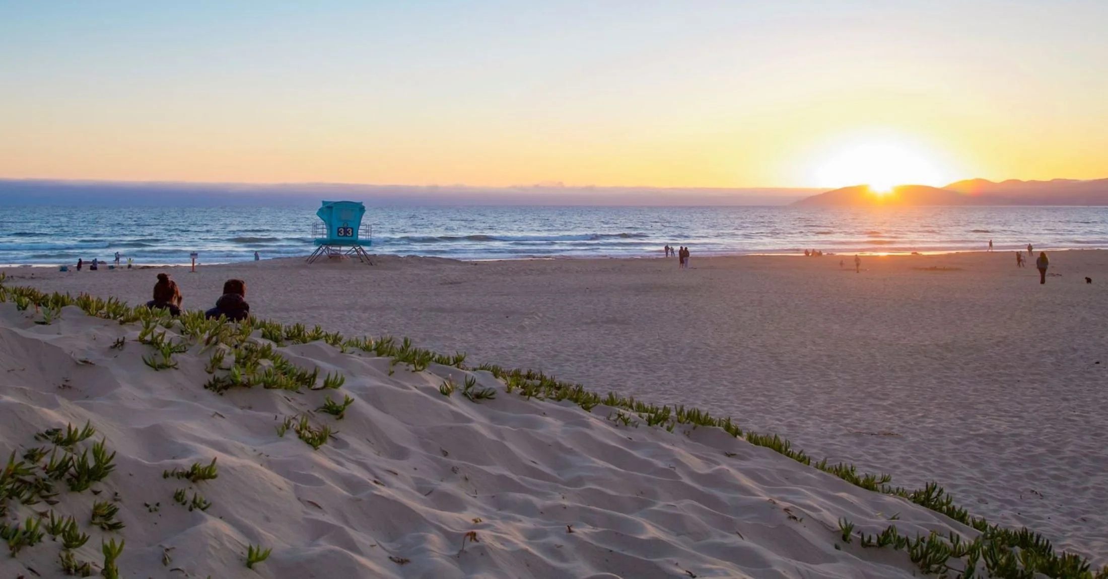 Aerial view of a residential neighborhood with golf course, trees, houses, and mountains in the background under a clear blue sky.