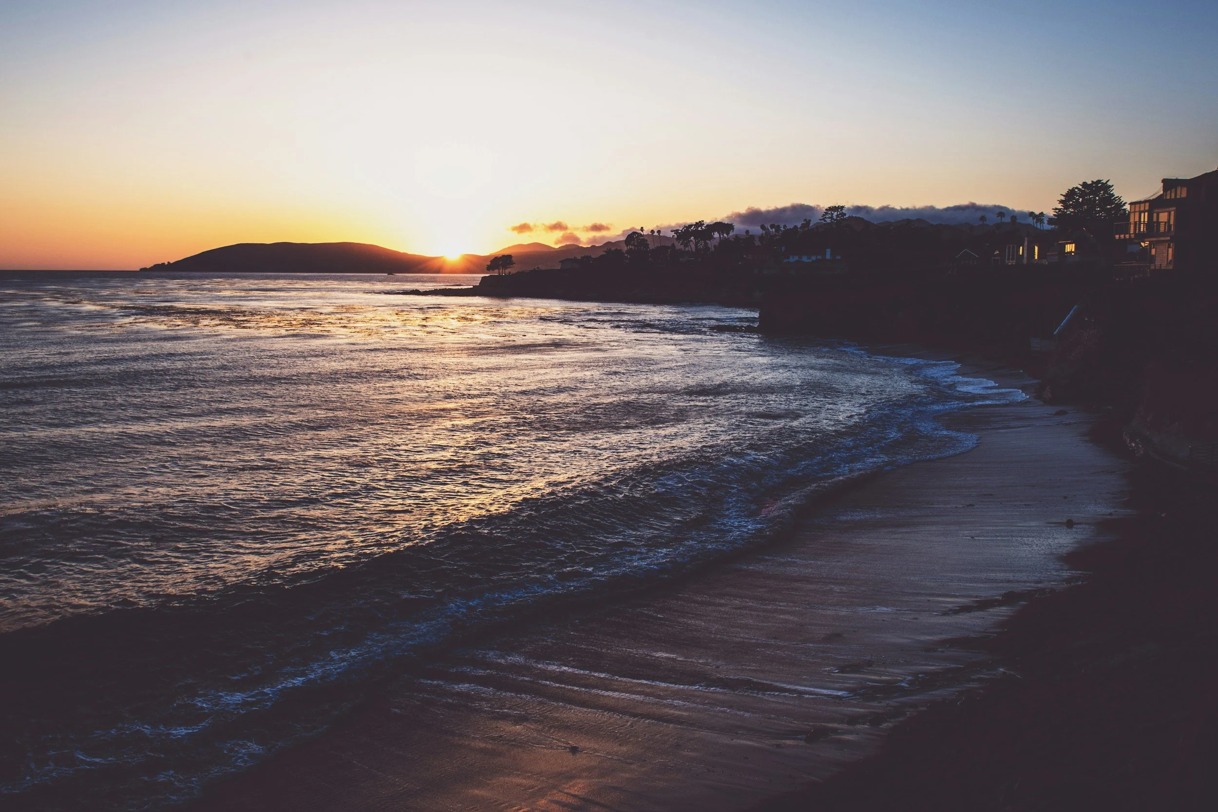 Aerial view of a coastal city with a hillside residential area, a parking lot, and the ocean in the background under a partly cloudy sky.