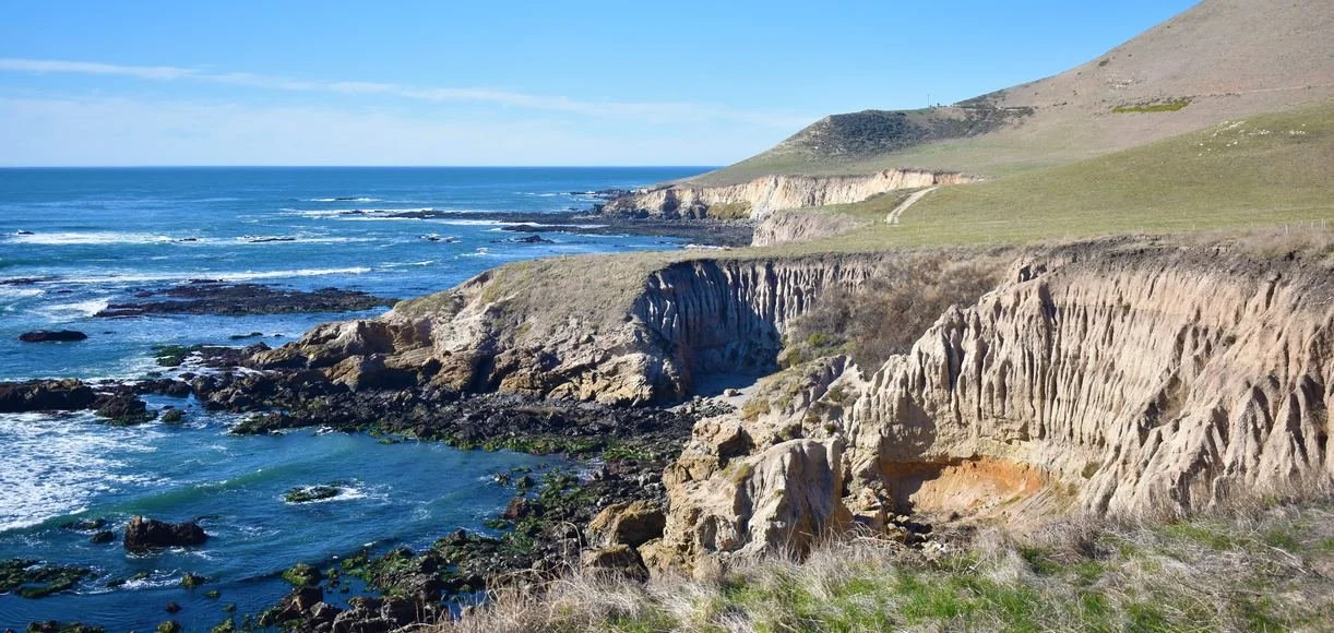 An aerial view of a small town with houses, trees, and streets, surrounded by rolling hills and mountains in the background.