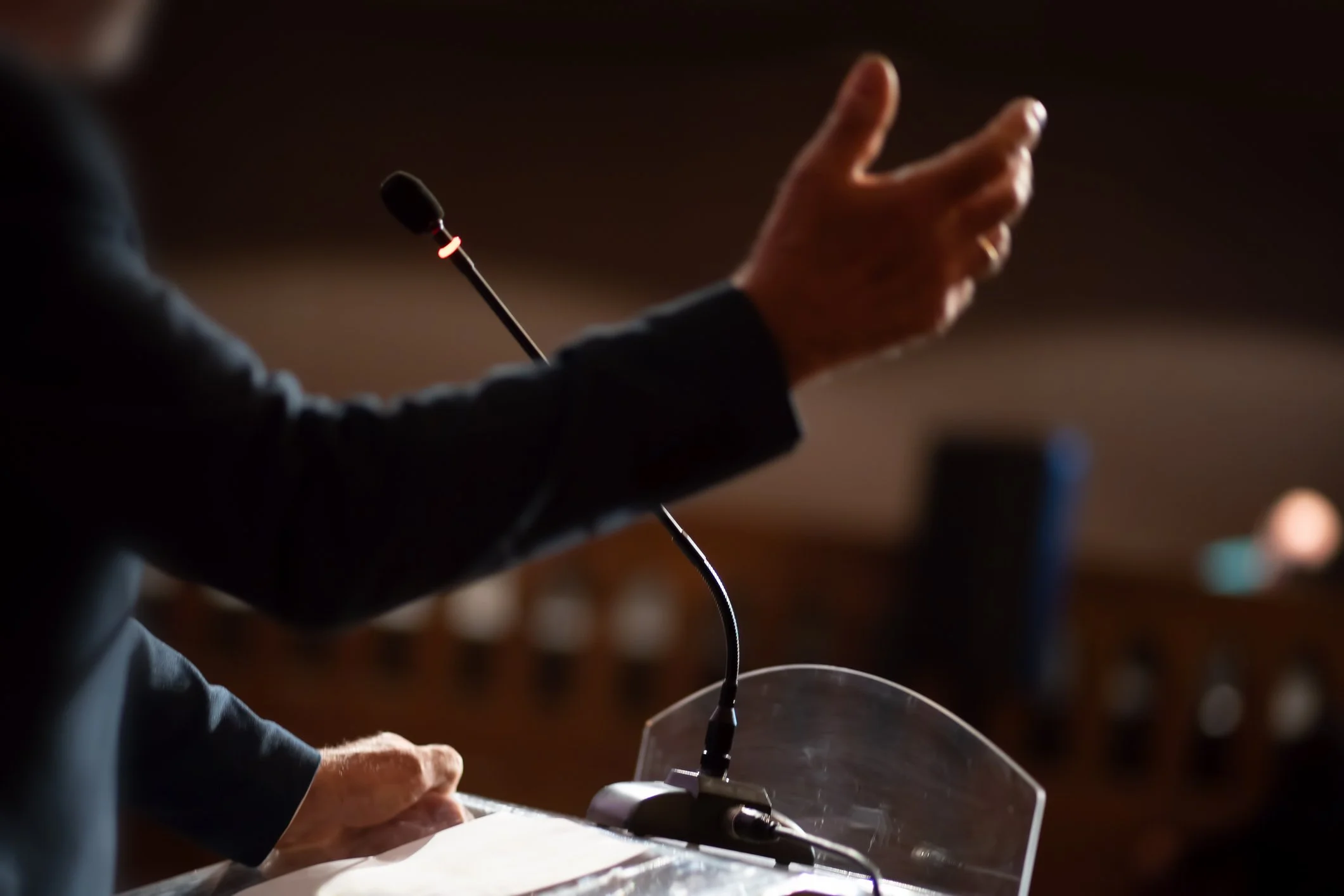 Person gesturing with their hand while speaking at a podium with a microphone in a dimly lit room.