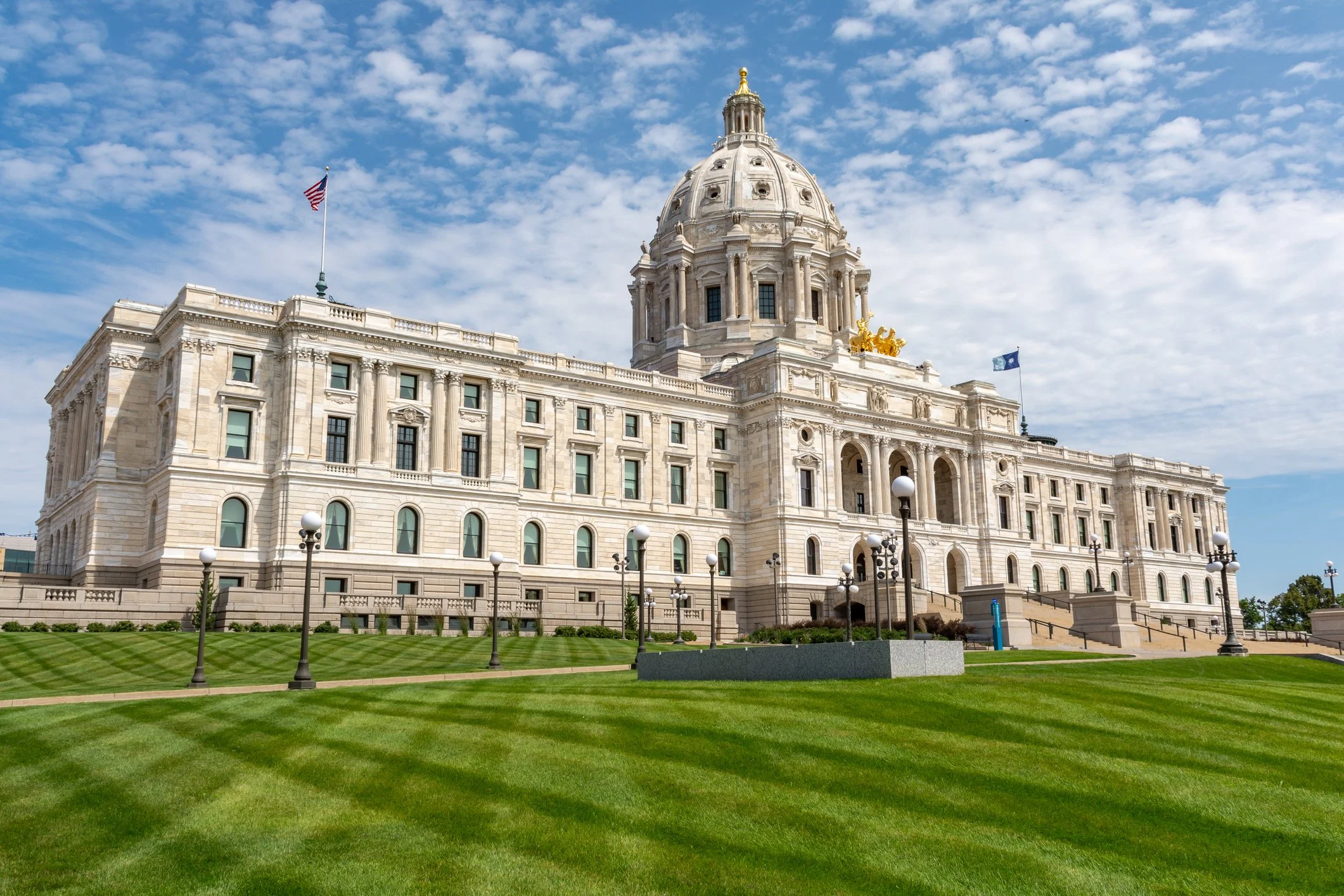 The image shows a large, historic, domed government building with white stone architecture, surrounded by a well-maintained green lawn and lamp posts under a partly cloudy sky.