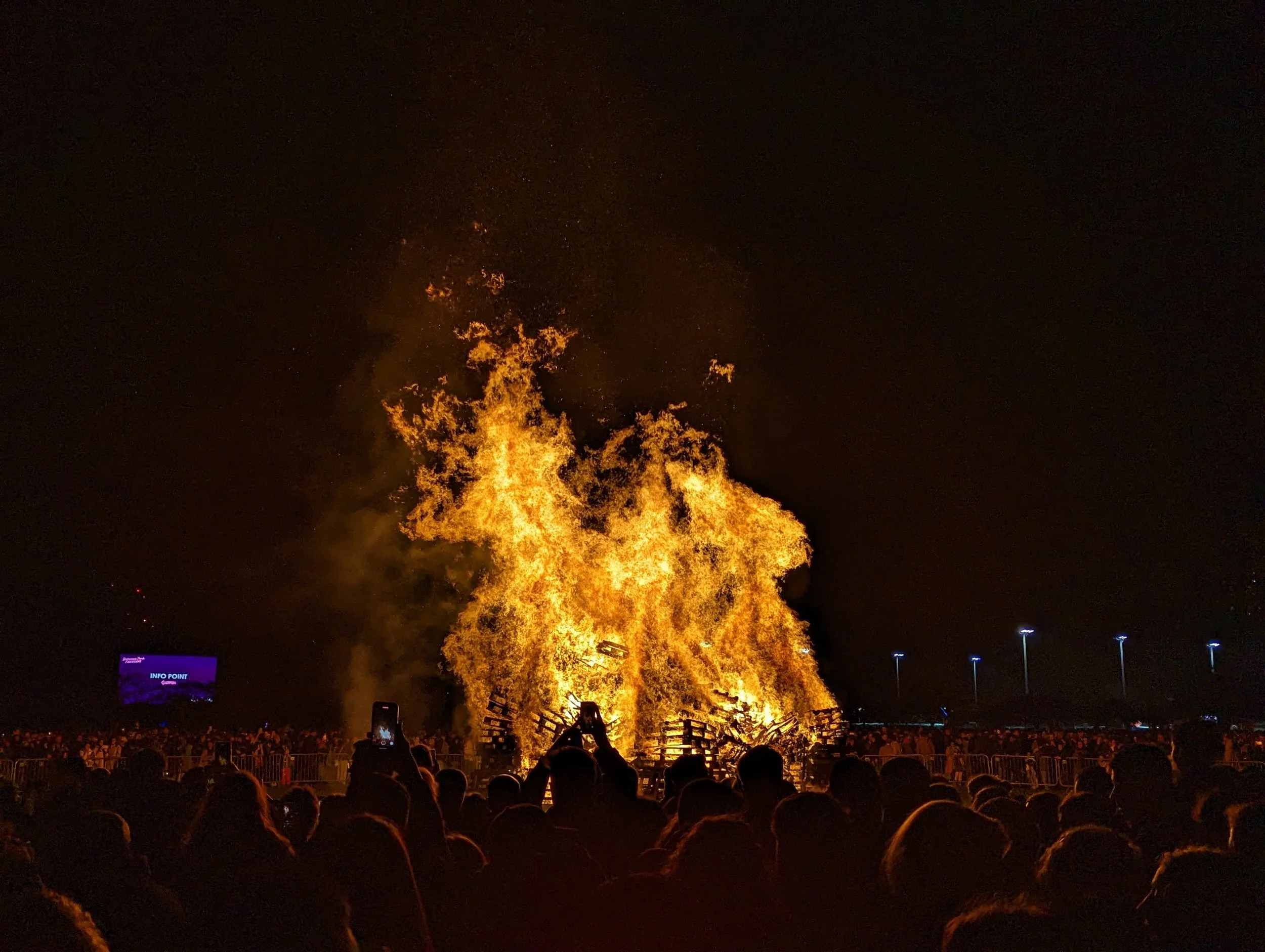 Large crowd of people watching a fireworks display at night, with bright flames and smoke rising into the dark sky, and a purple information sign in the foreground.