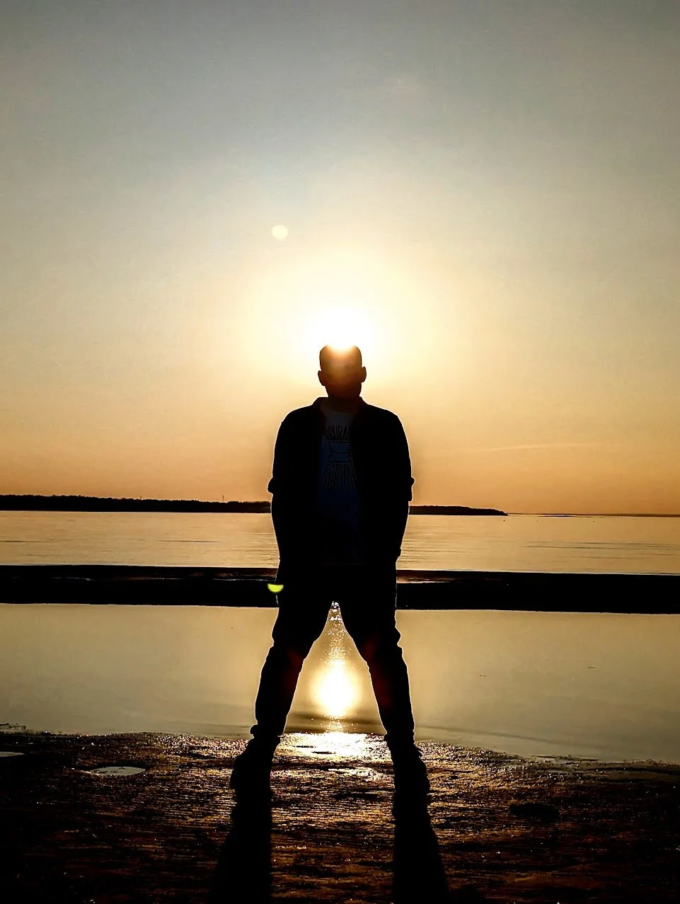 A person standing on a beach at sunset with their silhouette visible, facing water with the sun setting behind them and their shadow cast on the ground.