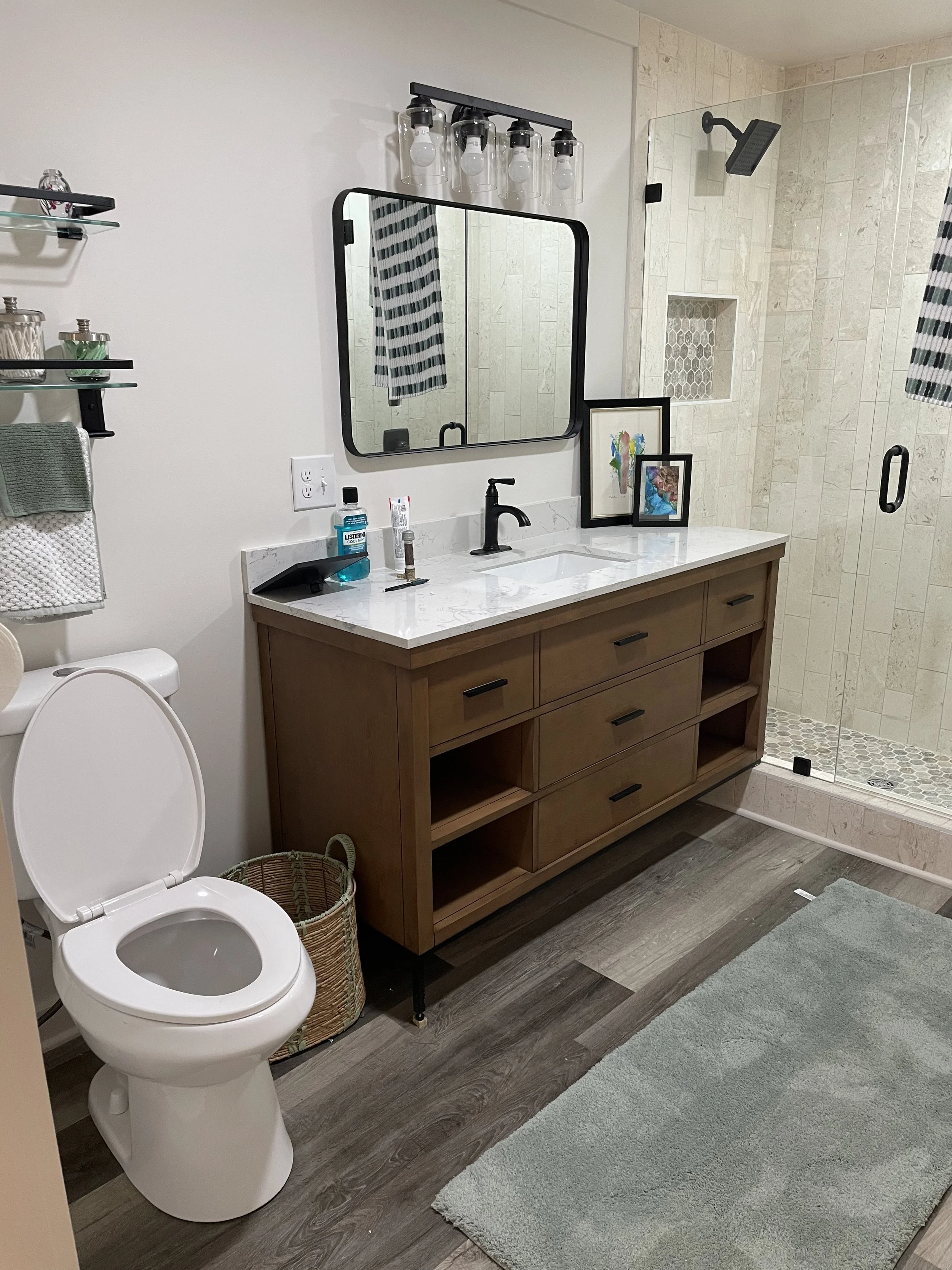 Modern bathroom with a white toilet, a wooden vanity with a white marble countertop, a black faucet, two framed artworks, a glass shower with a black sliding door, and glass shelves with toiletries.