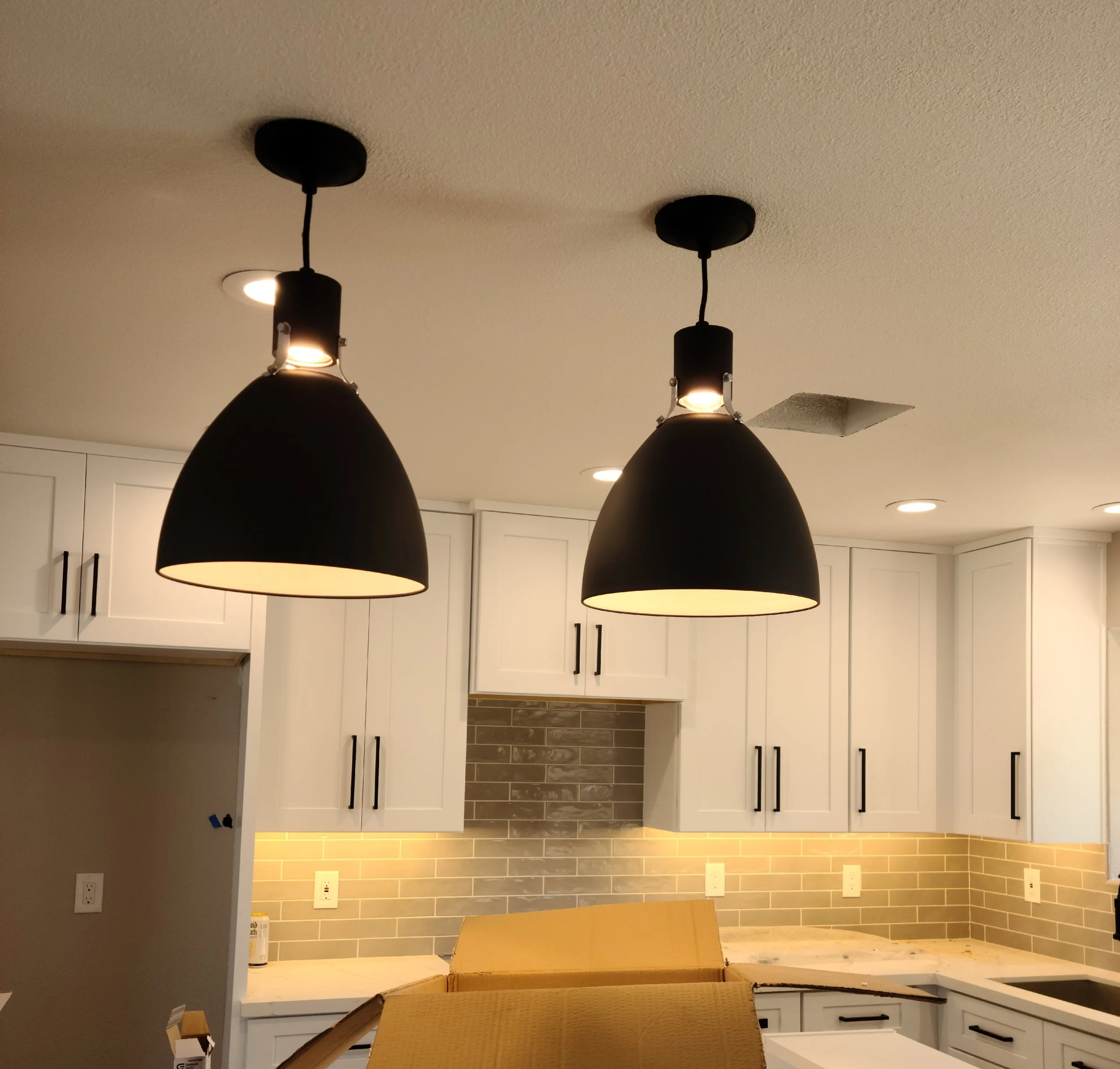 Two black pendant lights hanging over a kitchen with white cabinets, beige countertops, and a gray brick backsplash.
