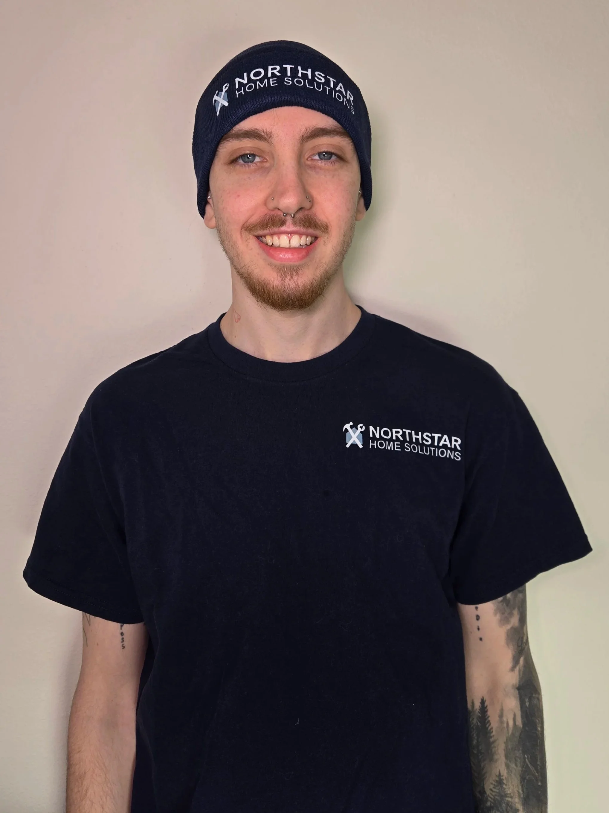 A young man with a beard and tattoos on his arm, smiling, wearing a black Northstar Home Solutions t-shirt and a navy blue beanie with the logo.