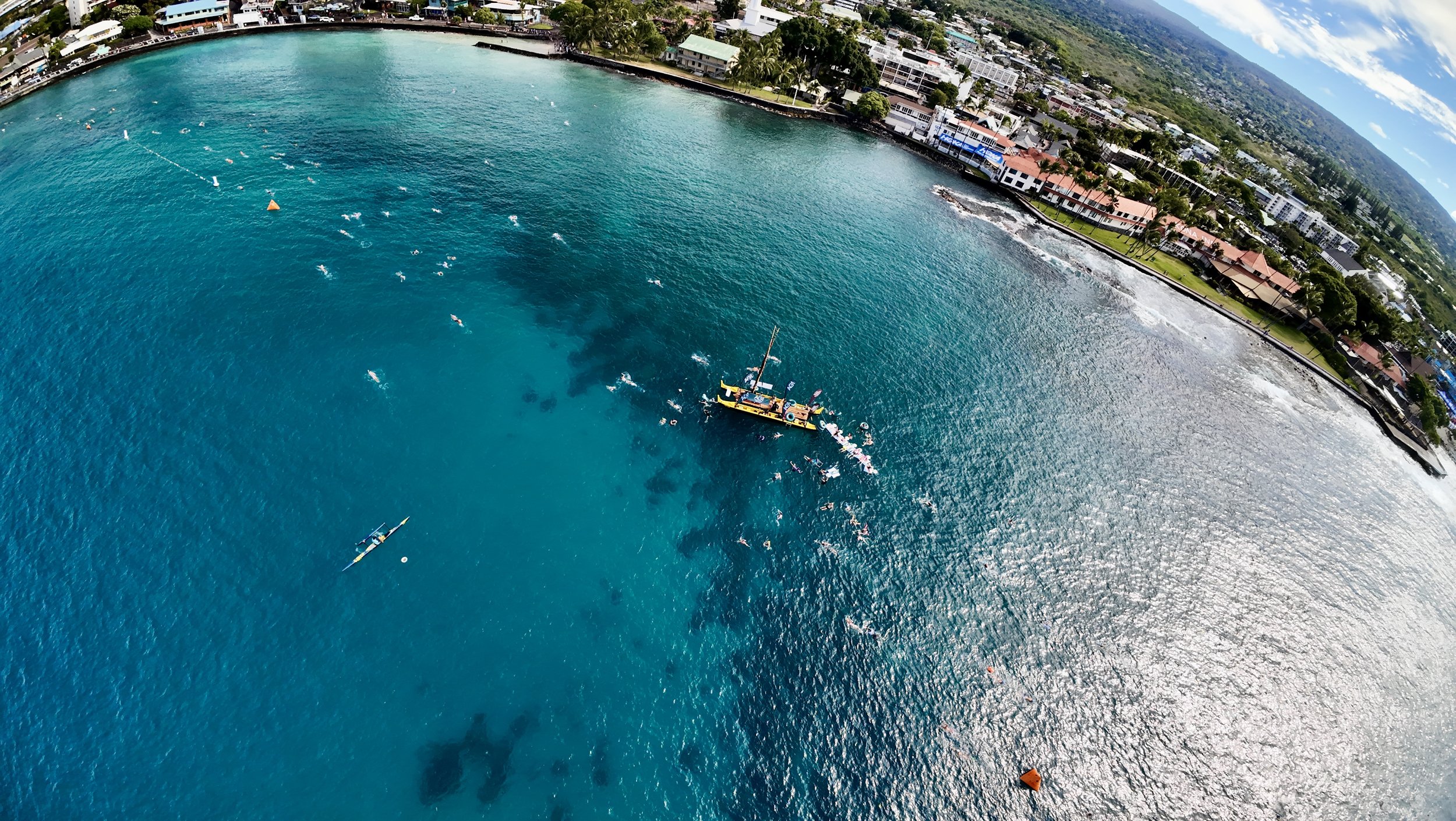 Aerial view of a coastal city with turquoise water, a boat, and swimmers. The shoreline features buildings and palm trees.