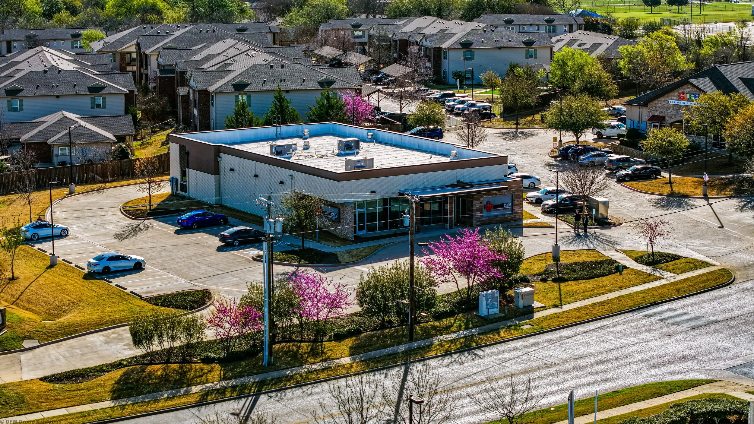 Aerial view of a small commercial building with a parking lot and surrounding residential area. The building is surrounded by green lawns and blooming trees. Several vehicles are parked, and nearby there are residential homes with roads and trees visible.