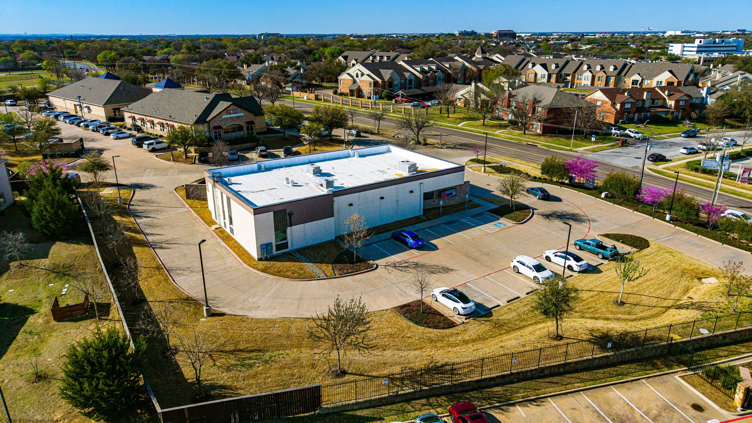 Aerial view of a small commercial plaza with a white-roofed building and several parked cars, surrounded by suburban houses and roads.