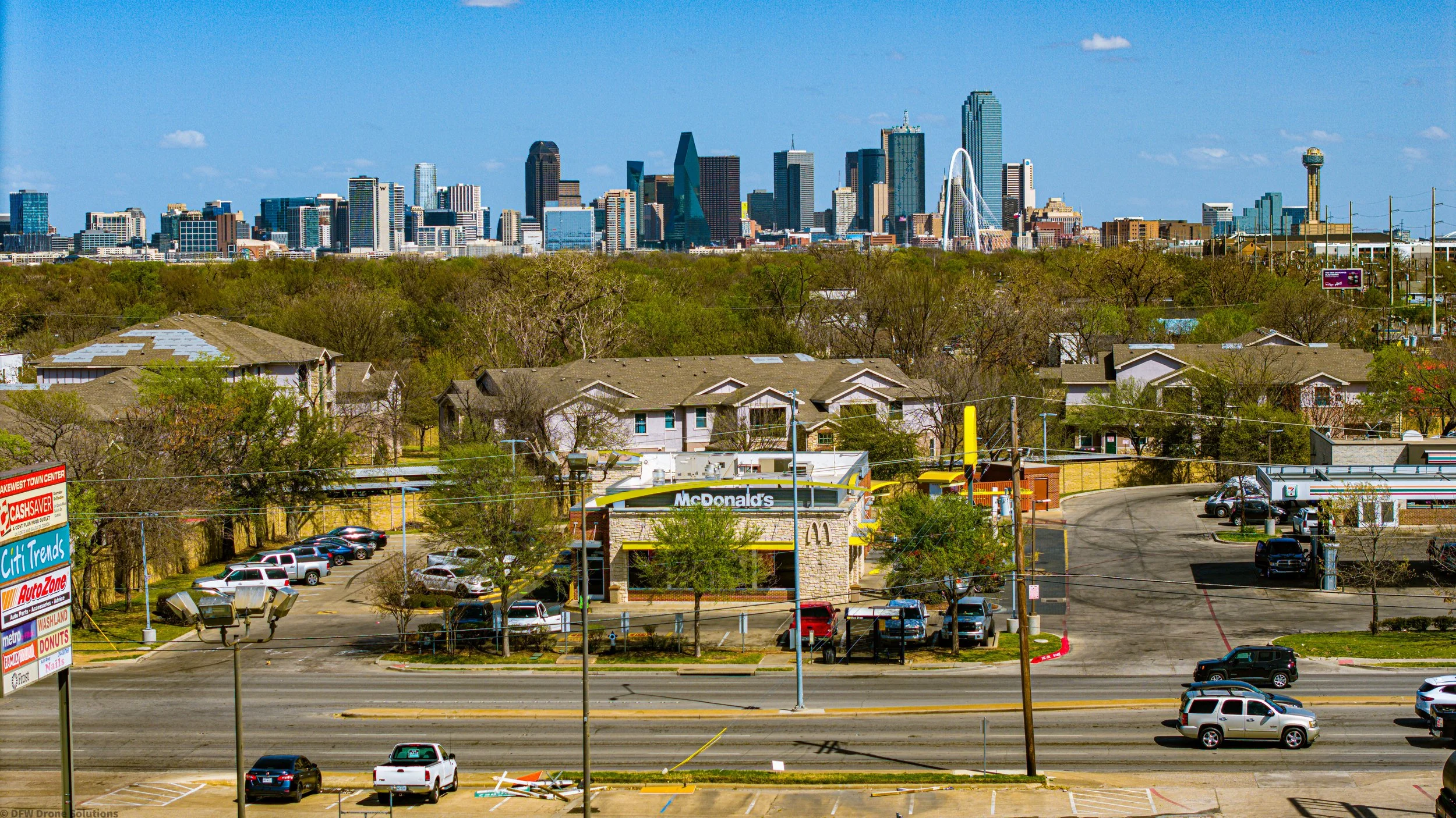 View of a city skyline with skyscrapers and a McDonald's in the foreground, surrounded by residential buildings and trees under a clear blue sky.